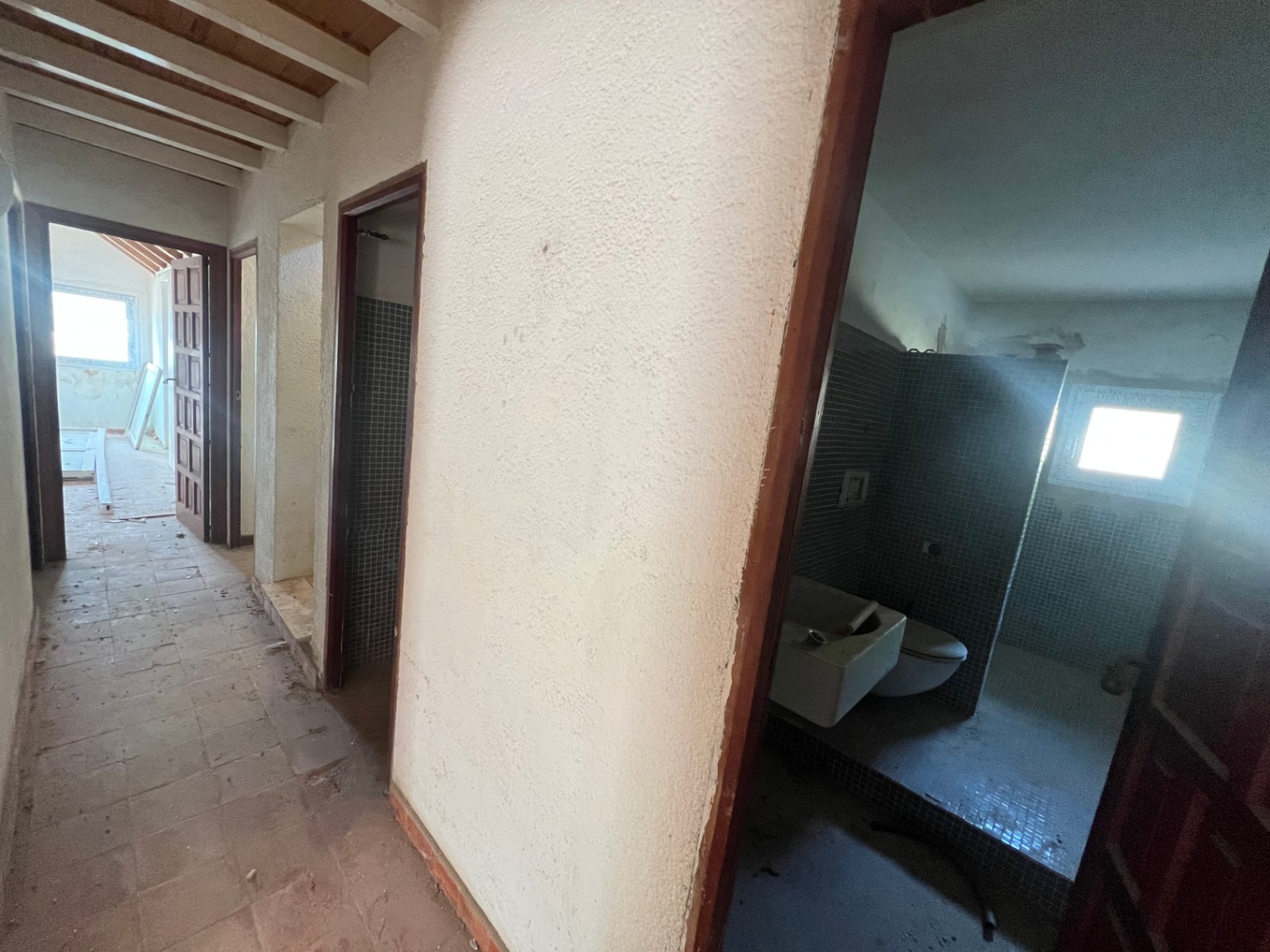 Binnengang met houten plafondbalken, terracotta vloertegels en toegang tot een badkamer met blauwe mozaïekbetegeling.