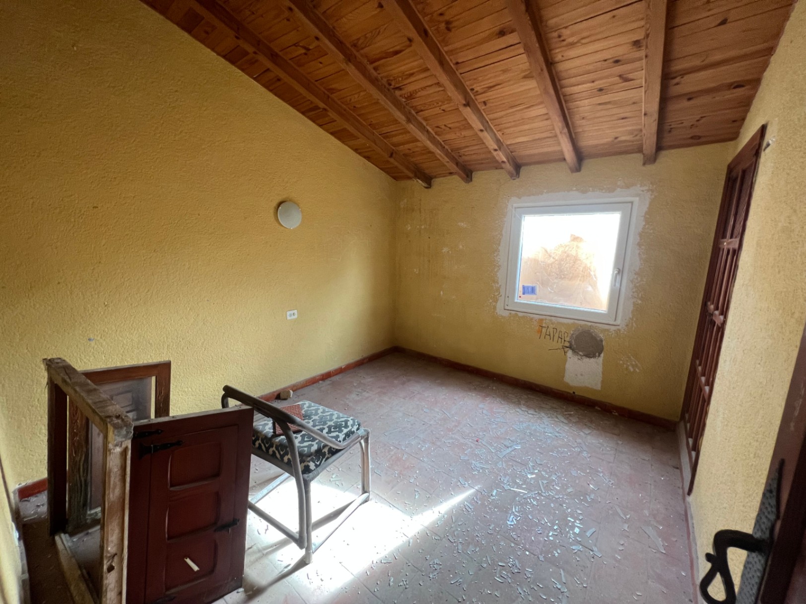 Binnenruimte met een schuin houten balkenplafond, terracotta vloertegels en gele muren. De kamer dient volledig gerenoveerd te worden en heeft natuurlijke lichtinval.