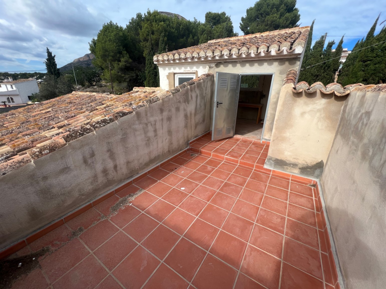 Bovenliggend terras met terracotta tegels en toegang tot een klein bijgebouw met een pannendak, gelegen in een groene omgeving nabij de Montgó.