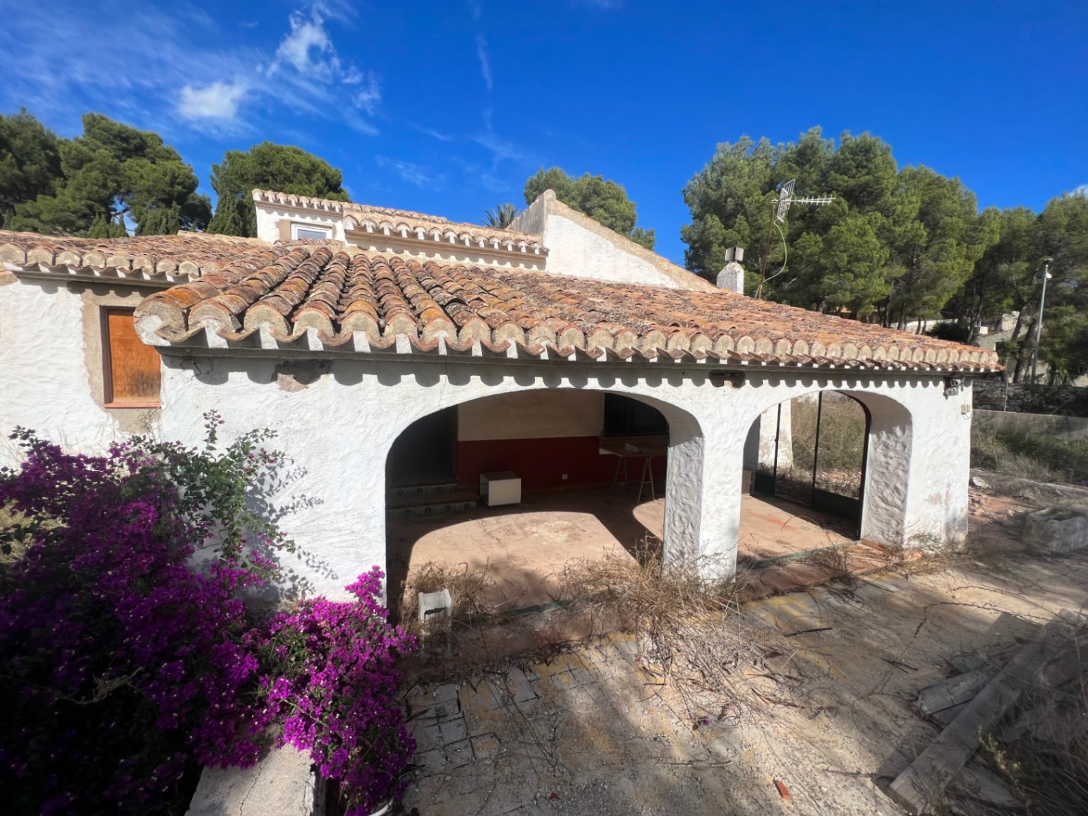 Buitenaanzicht van een traditionele naya met witte bogen en terracotta dakpannen. De mediterrane architectuur is omgeven door pijnbomen en paarse bougainville.