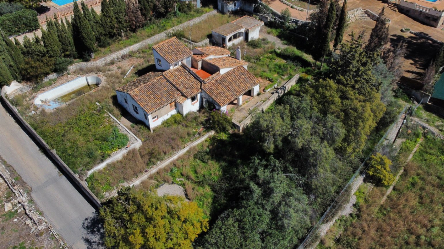 Luchtfoto van de villa met diverse pannendaken, het uitgestrekte beboste perceel en het te renoveren zwembad, gelegen in de wijk Montgó.