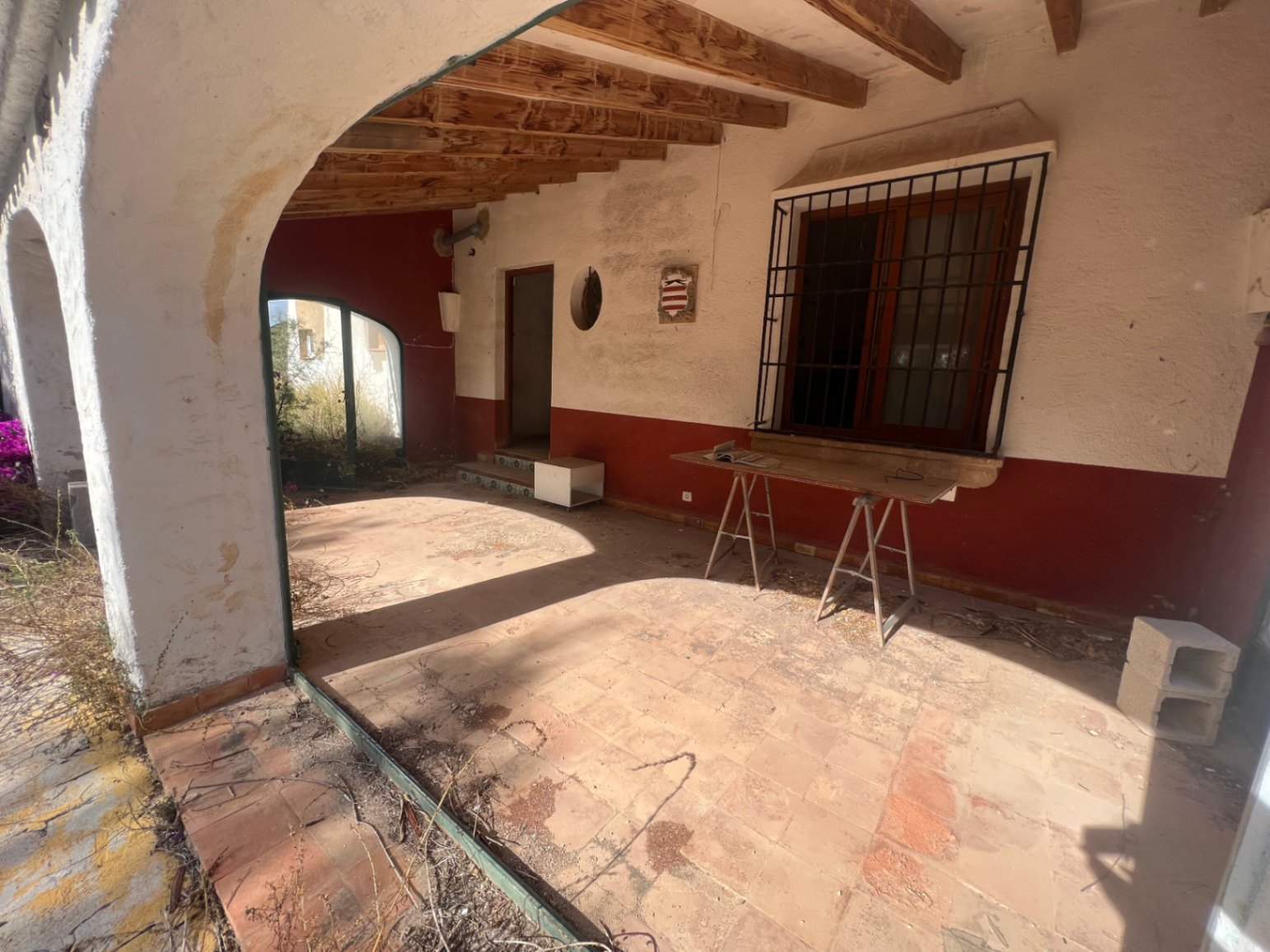 Overdekt terras met houten plafondbalken, terracotta vloertegels en traditionele bogen. De ruimte vereist een volledige renovatie om de mediterrane stijl te herstellen.