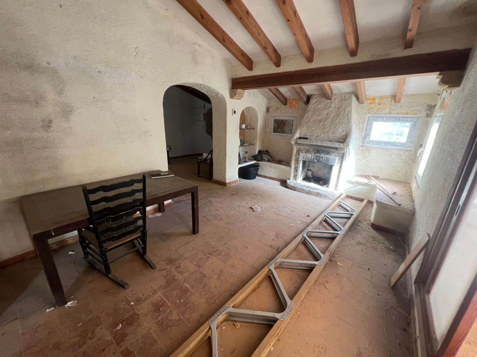 Rustieke woonkamer met houten plafondbalken, authentieke terracotta vloertegels en een ingebouwde open haard. De ruimte is voorzien van schuine daken en boogdoorgangen.