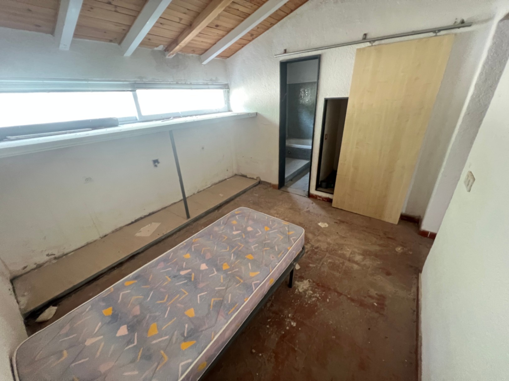 Slaapkamer met schuin plafond met houten balken, horizontaal raam en een schuifdeur die toegang biedt tot een aangrenzende badkamer met donkere tegels.