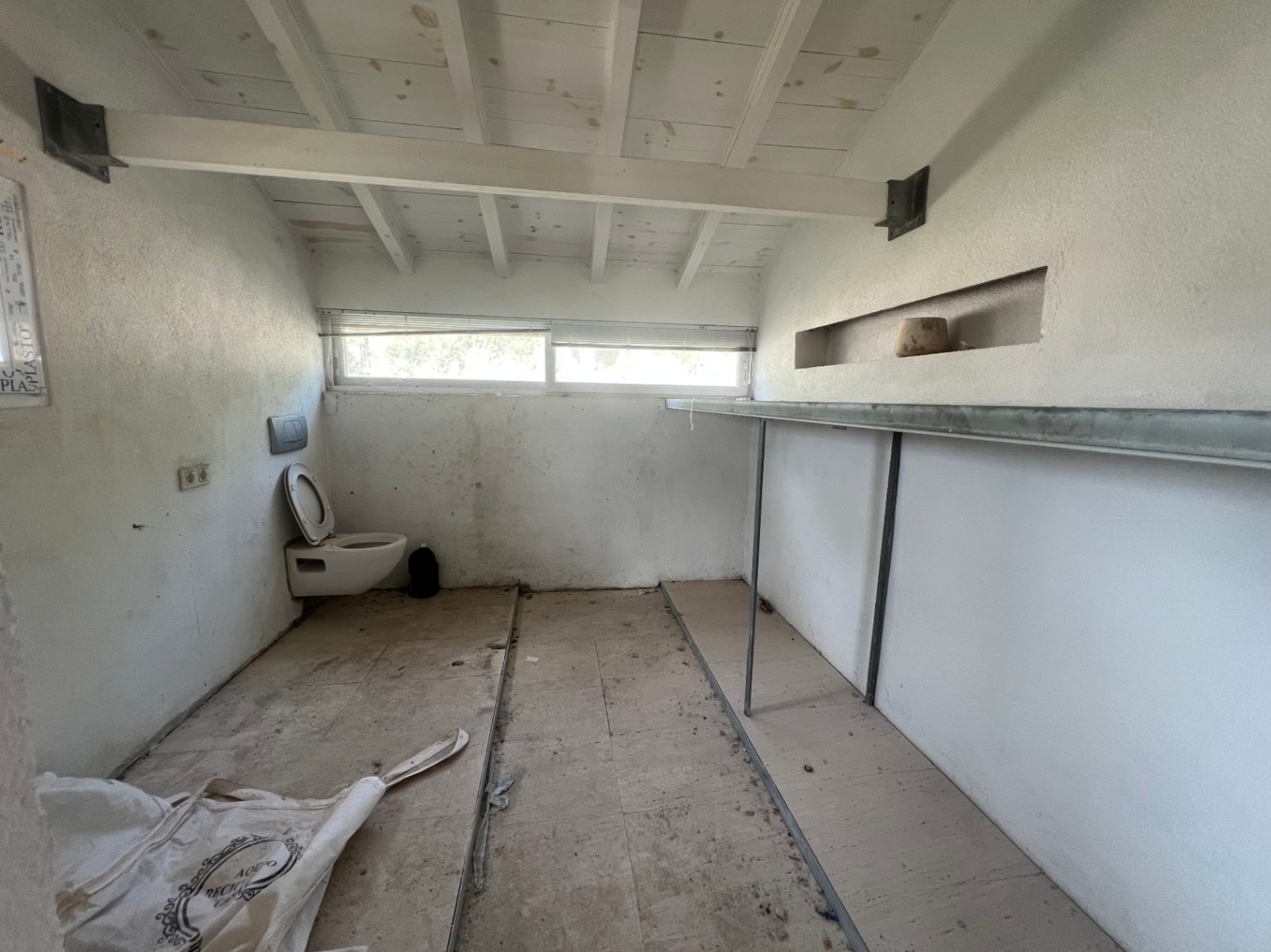 Te renoveren binnenruimte met wit houten balkenplafond, hangend toilet en vaste schappen. Een horizontaal raam zorgt voor natuurlijke lichtinval.