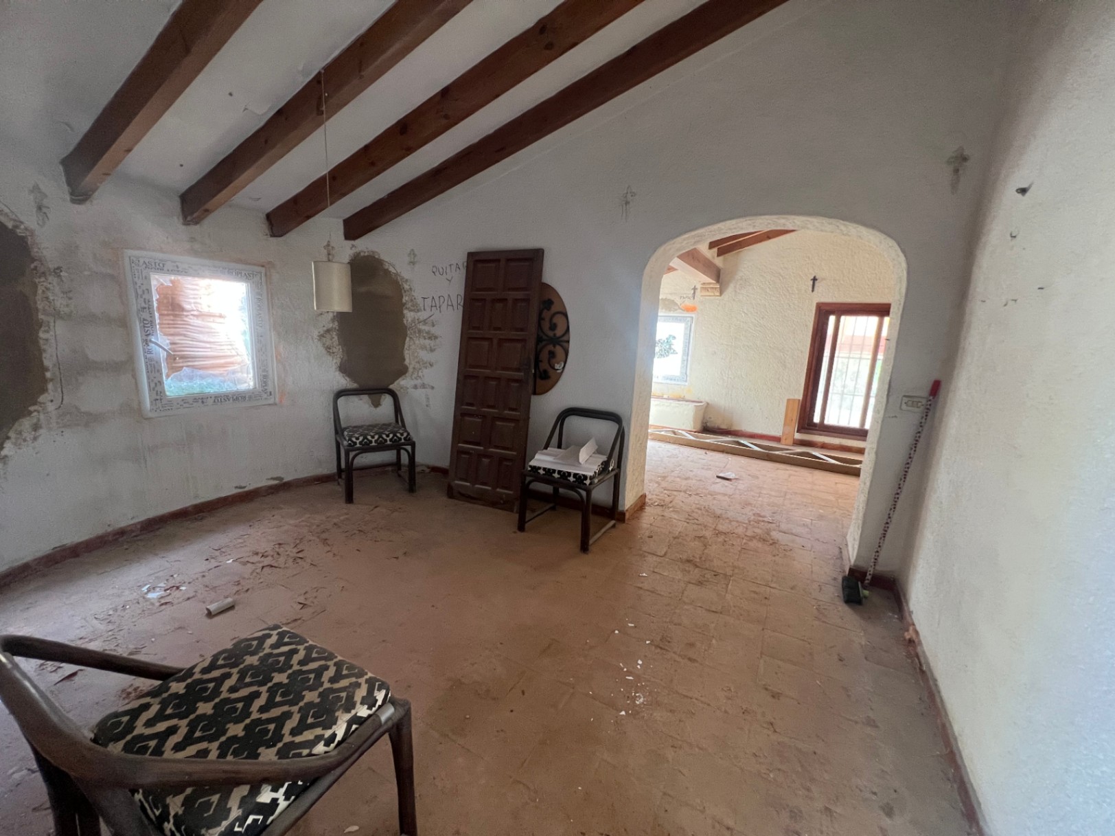 Te renoveren binnenruimte met zichtbare houten plafondbalken, terracotta tegelvloer en een karakteristieke boogdoorgang naar de volgende kamer.