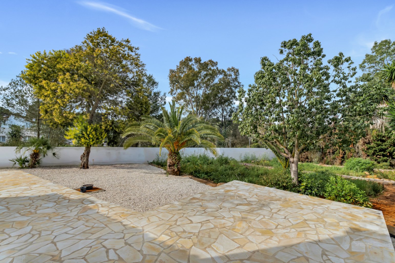 Buitenterras met onregelmatige natuurstenen bestrating, uitkijkend op een mediterrane tuin met palmbomen, grindpartijen en volwassen beplanting onder een heldere hemel.