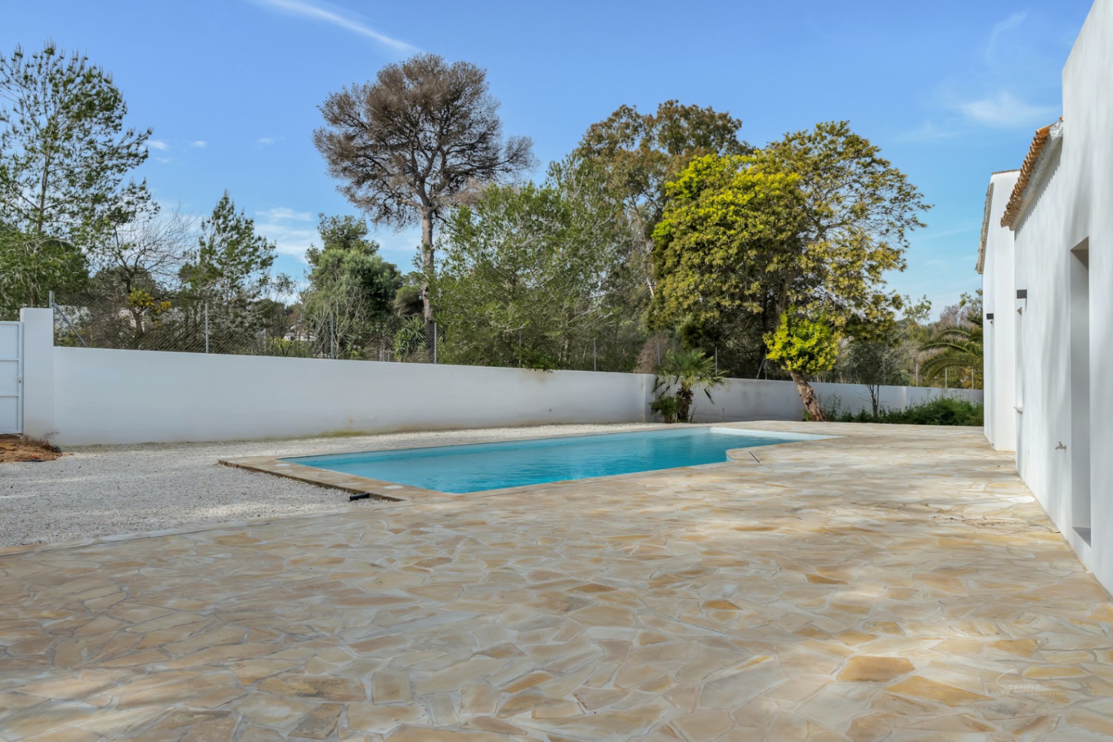 Buitenzwembad omgeven door een ruim terras van natuursteen en witte muren, gelegen in een tuin met volwassen mediterrane bomen.