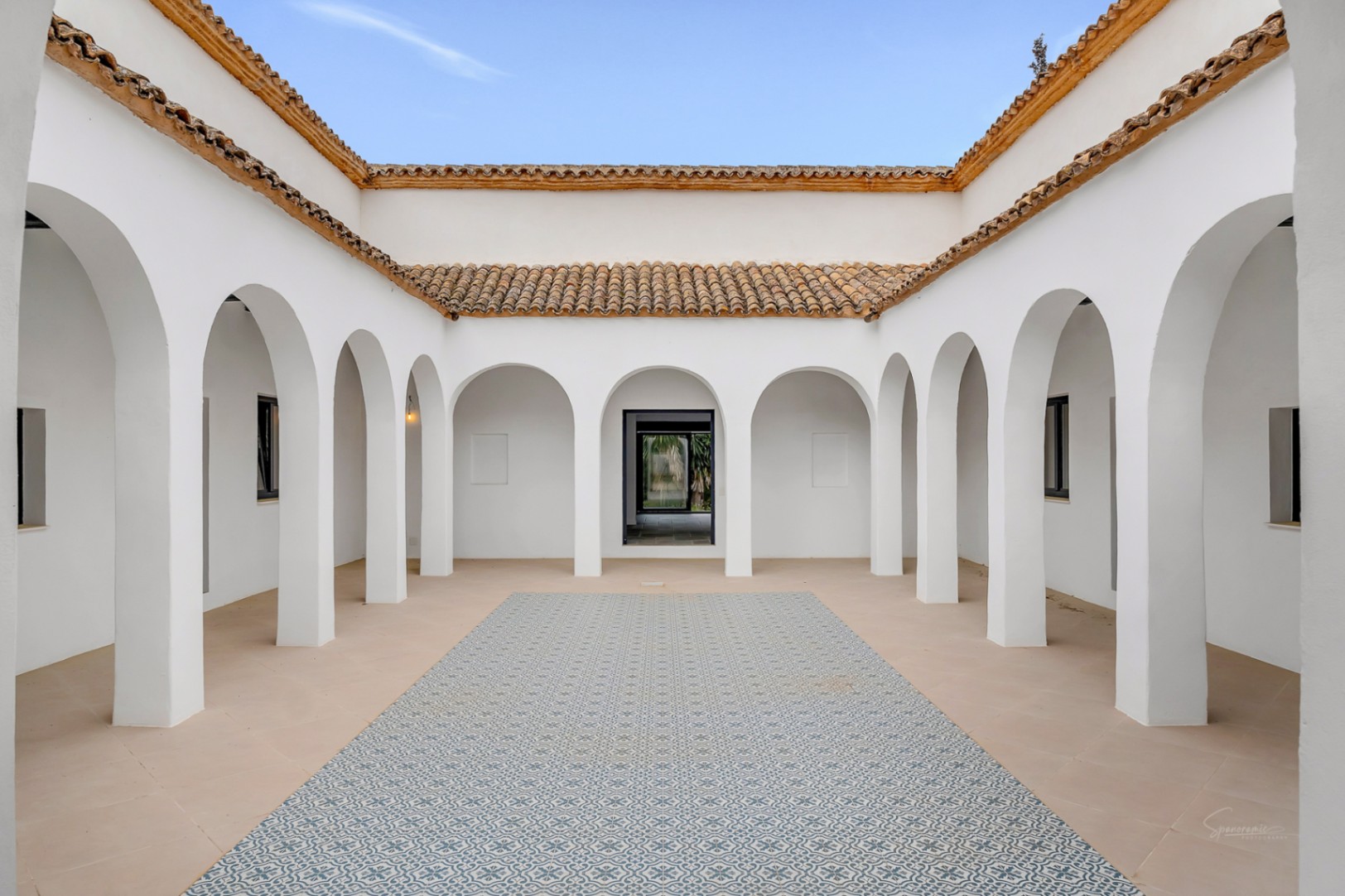 Centrale mediterrane binnenplaats met symmetrische witte bogen, traditionele terracotta dakpannen en een decoratieve blauwe patroontegelvloer.