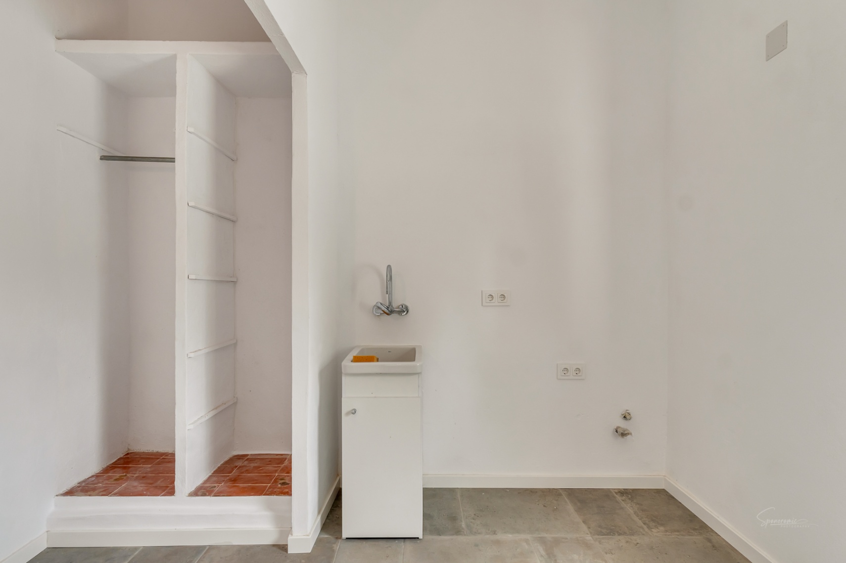 Functionele bijkeuken met een compacte wastafel, stopcontacten en een ingebouwde nishoek voor opslag met terracotta tegels en een stenen vloer.