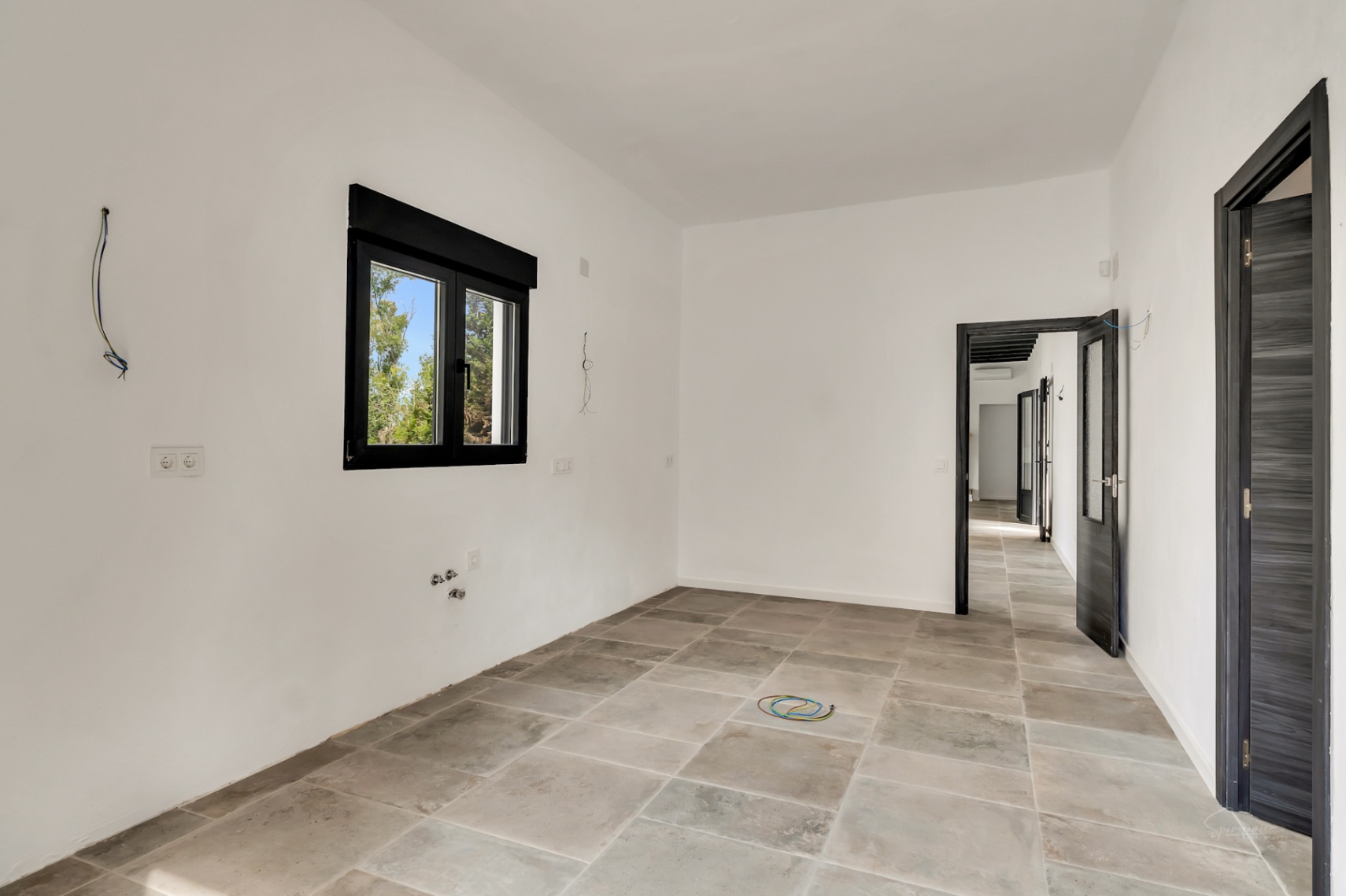 Kamer in renovatie met vloertegels in steenlook, witte muren en donkere kozijnen. Voorzien van zichtbare aansluitingen voor water en elektriciteit.