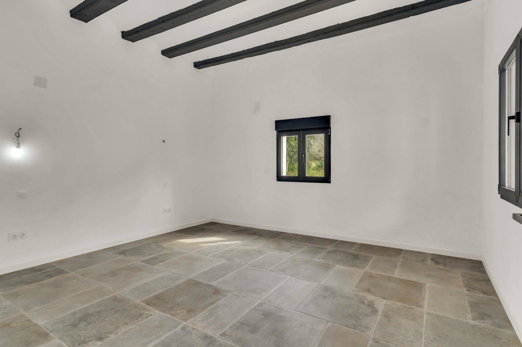 Lichte, lege kamer met natuurstenen vloertegels, donkere houten plafondbalken en zwart omrande ramen die zorgen voor een modern contrast met de witte muren.