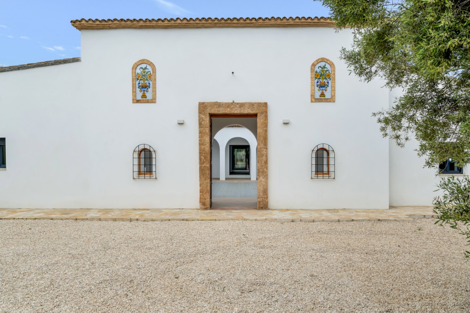 Mediterrane gevel met witte muren, een entree in Tosca-natuursteen en decoratieve keramische tegels. De centrale doorgang biedt zicht op de binnenplaats met bogen.