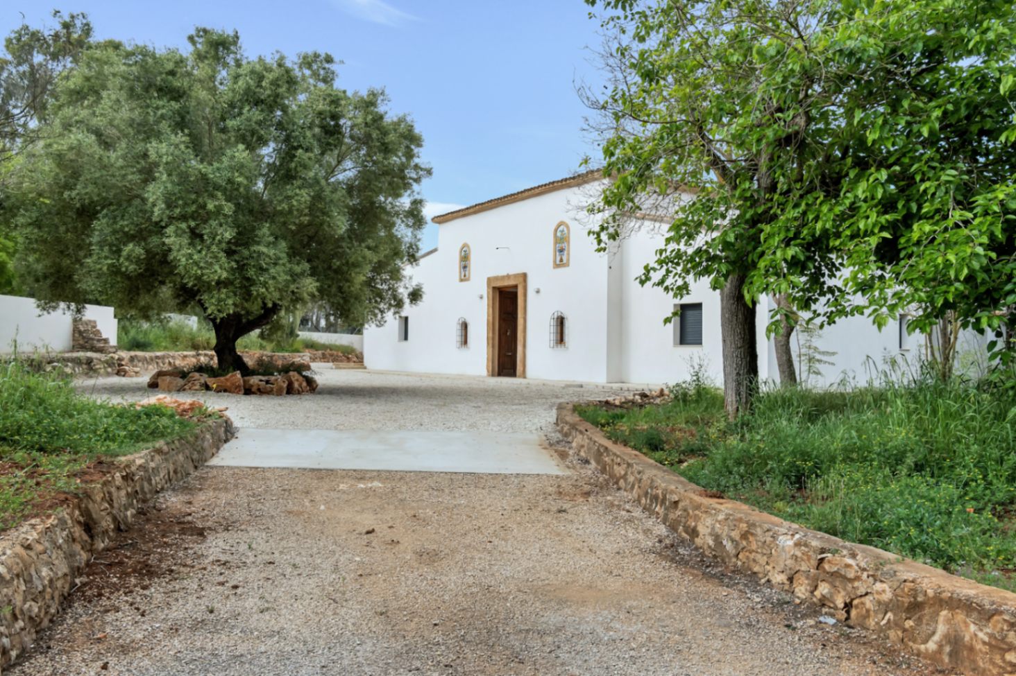 Mediterrane gevel met witte stucwerkmuren, Tosca-stenen deuromlijstingen en een grindpad omzoomd door natuurstenen muurtjes en volwassen olijfbomen.
