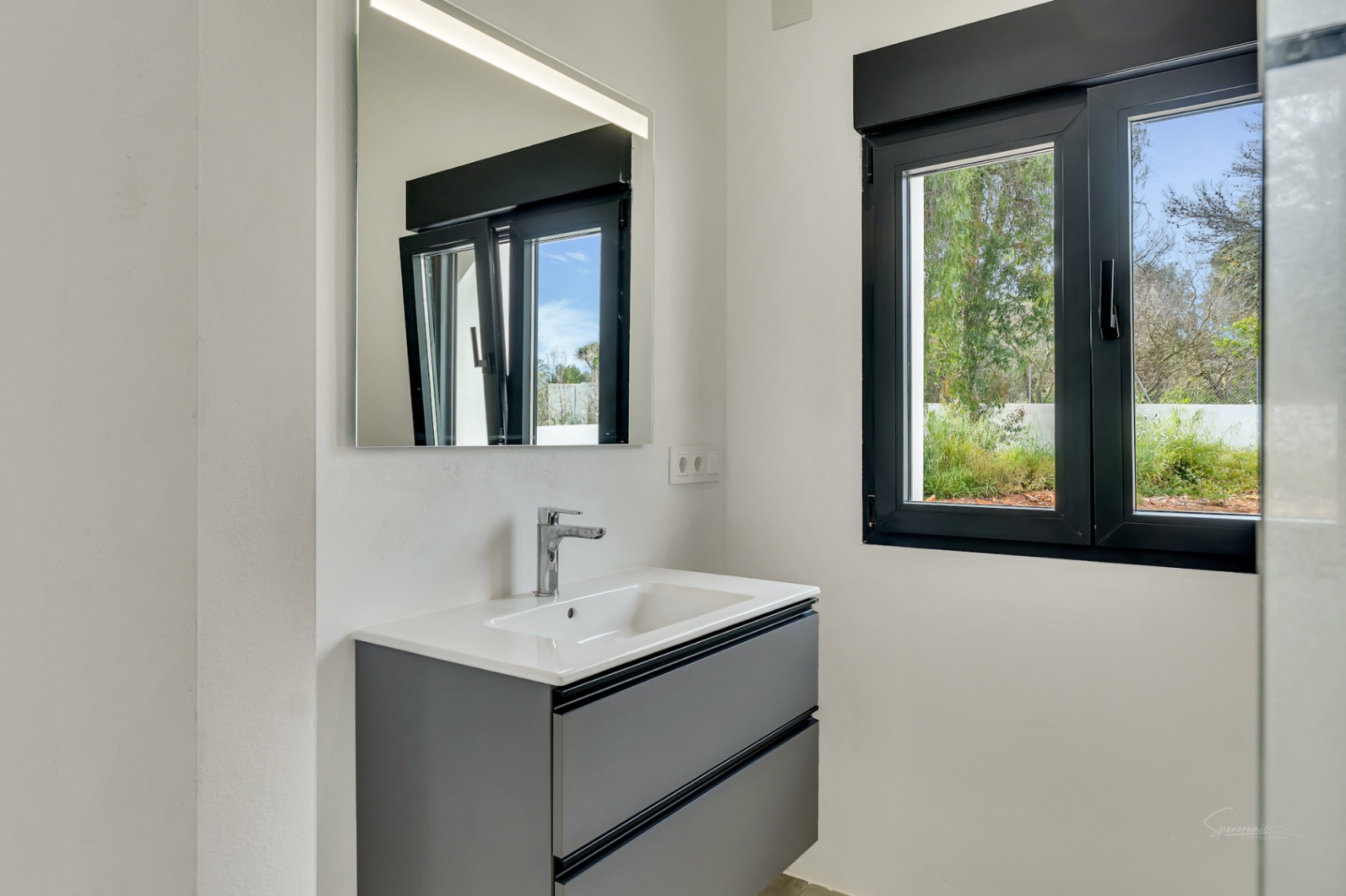 Moderne badkamer met een antraciet zwevend wastafelmeubel, verlichte spiegel en een zwart raamkozijn met uitzicht op de tuin.