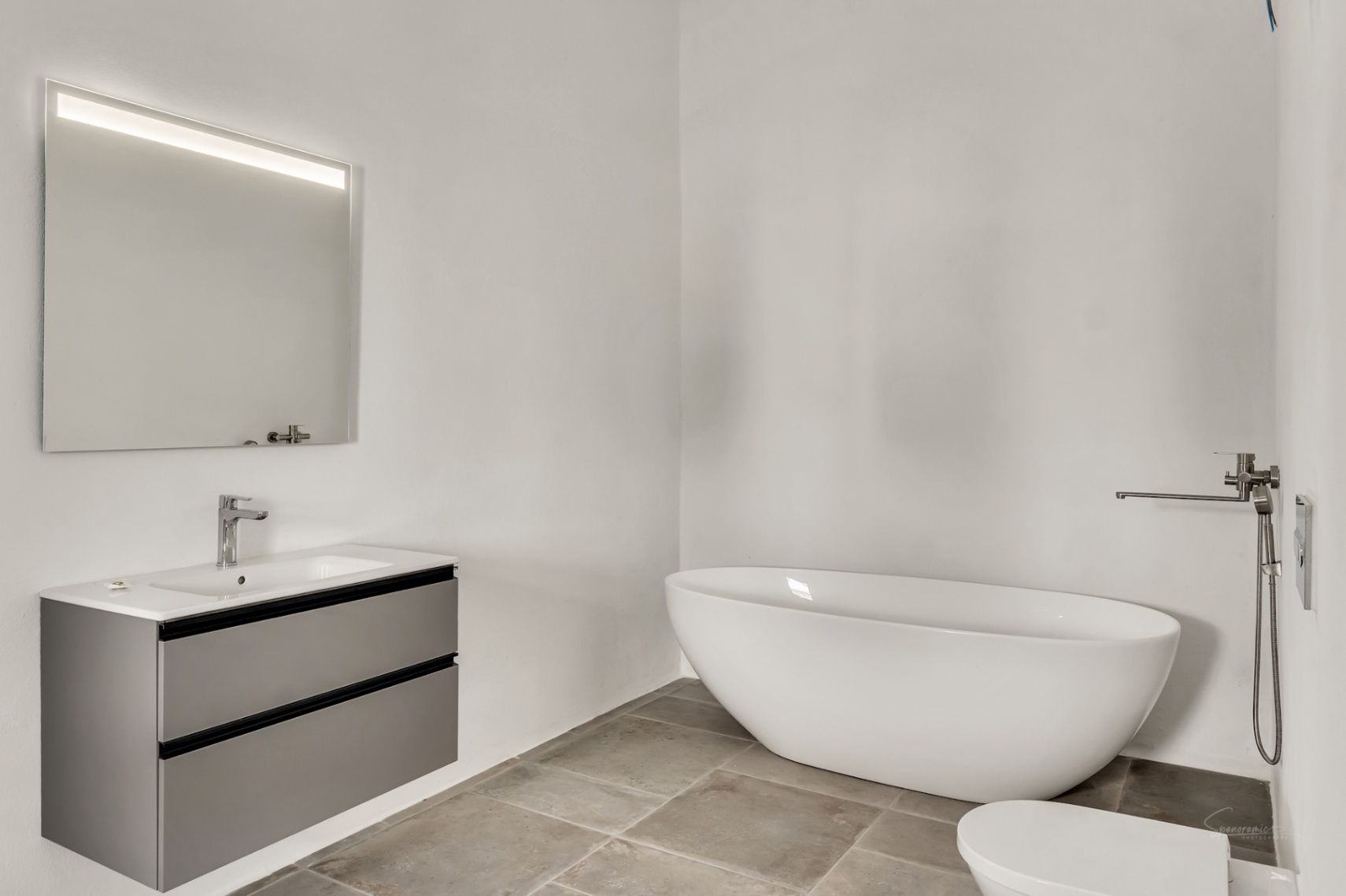 Moderne badkamer met een vrijstaand ovaal bad, een zwevend grijs wastafelmeubel en natuurstenen vloertegels tegen strakke witte wanden.