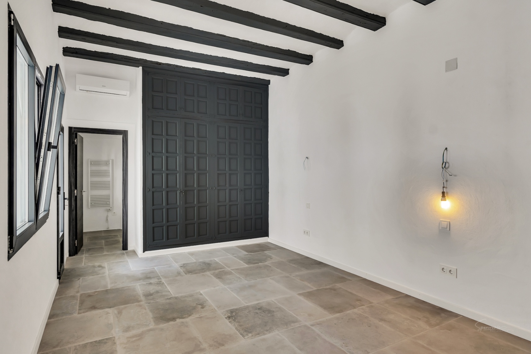 Slaapkamer met donkere houten balken, natuurstenen vloer en een grote inbouwkast met zwarte paneeldeuren. Voorzien van airconditioning en witte stucwerkwanden.