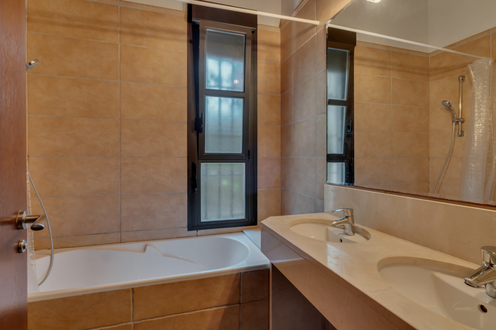 Badkamer met dubbele wastafel van natuursteen, ingebouwd ligbad en aardetinten wandtegels bij een raam met een donker profiel.