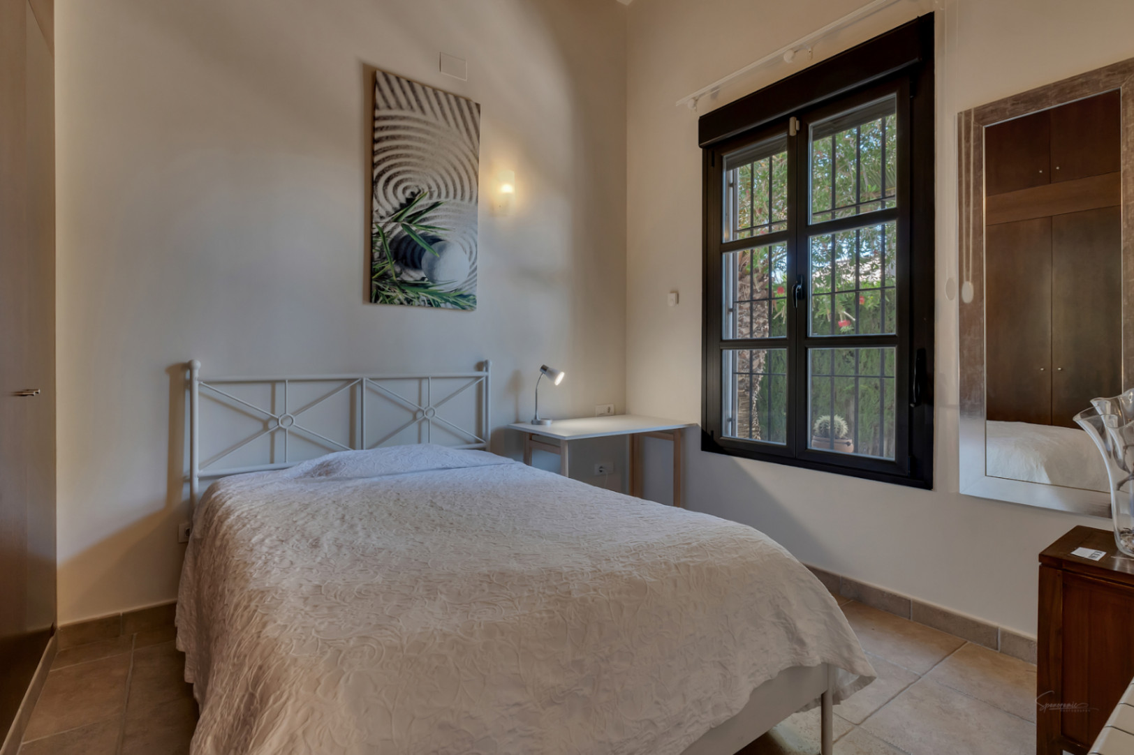 Lichte slaapkamer met wit metalen bedframe, bureau en raam met donker kozijn dat uitkijkt op de tuin. Voorzien van een tegelvloer en inbouwkasten.