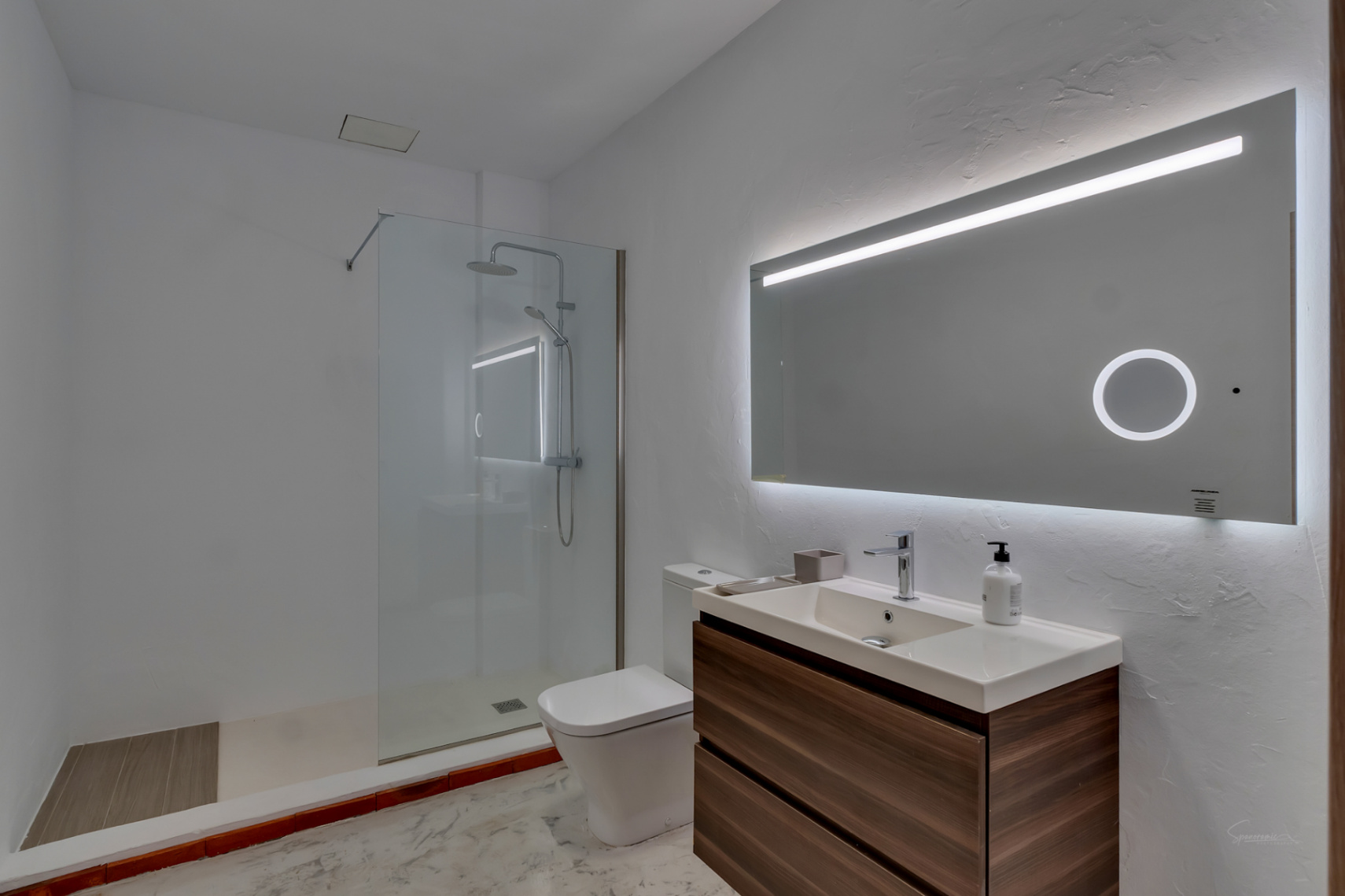 Moderne badkamer met een donker houten wastafelmeubel, LED-verlichte spiegel en inloopdouche. Voorzien van een glazen wand en een strakke, witte afwerking.