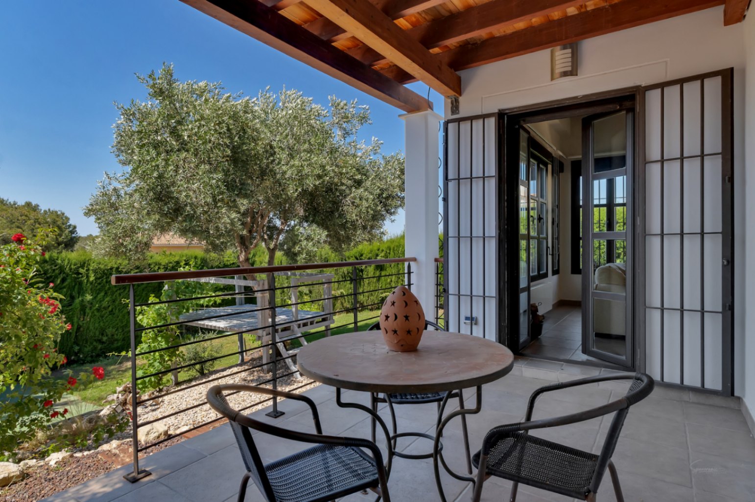 Overdekt terras met zichtbare houten balken, buitenmeubilair en uitzicht op de mediterrane tuin met een karakteristieke olijfboom.