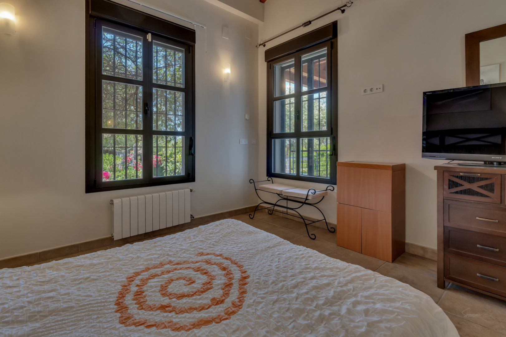 Slaapkamer met donkere raamkozijnen en tralies, uitzicht op de tuin, wandradiator, smeedijzeren bankje en houten ladekast met televisie.