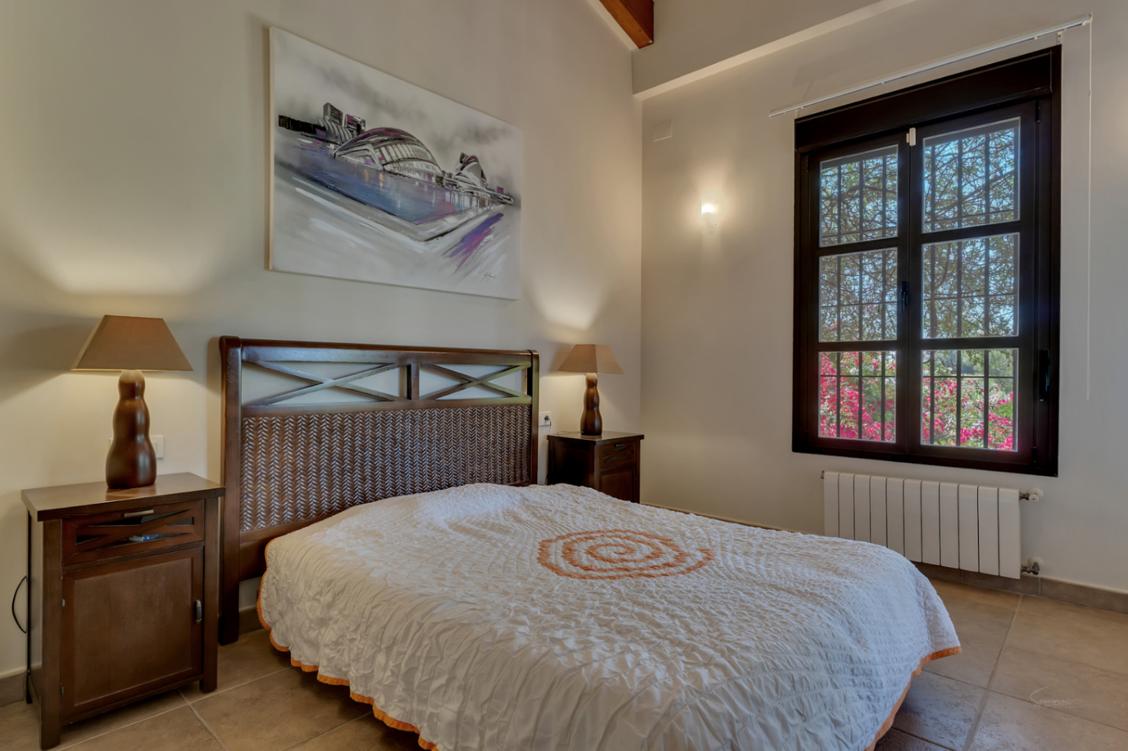 Slaapkamer met donkerhouten bed, gevlochten hoofdbord en zichtbare plafondbalken. Voorzien van een tegelvloer en houten kozijnen met uitzicht op de tuin.