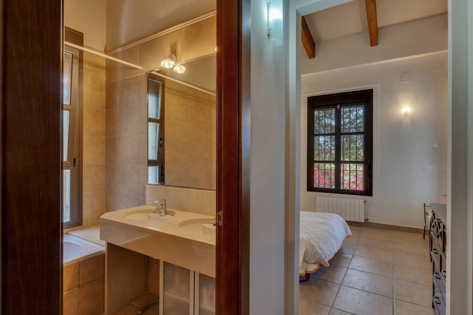 Uitzicht vanuit de gang op een badkamer met dubbele stenen wastafel en een slaapkamer met houten balkenplafond en uitzicht op de tuin.