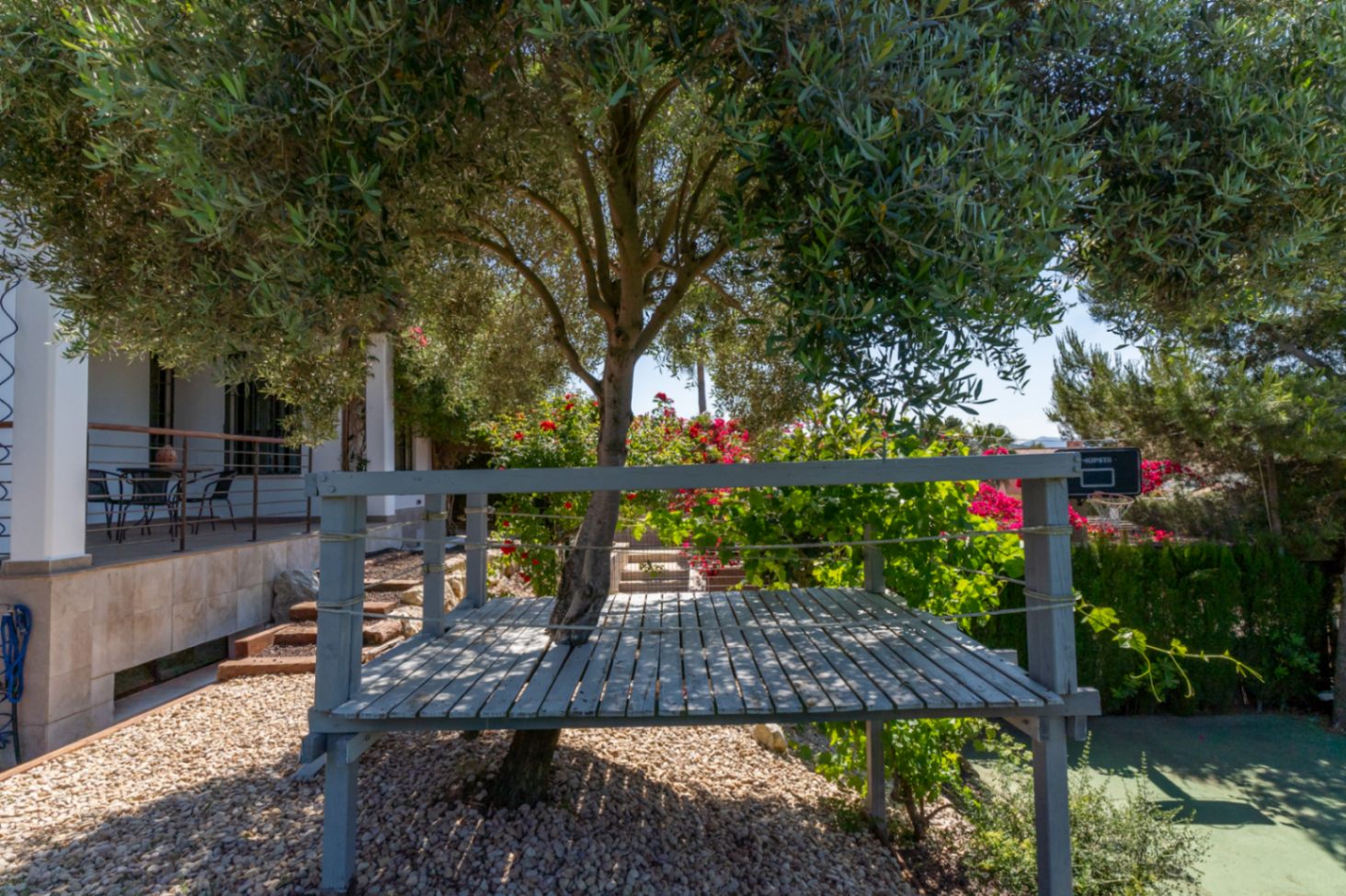 Verhoogd grijs houten platform rondom een olijfboom, gelegen in een mediterrane tuin met grind en bloeiende struiken.