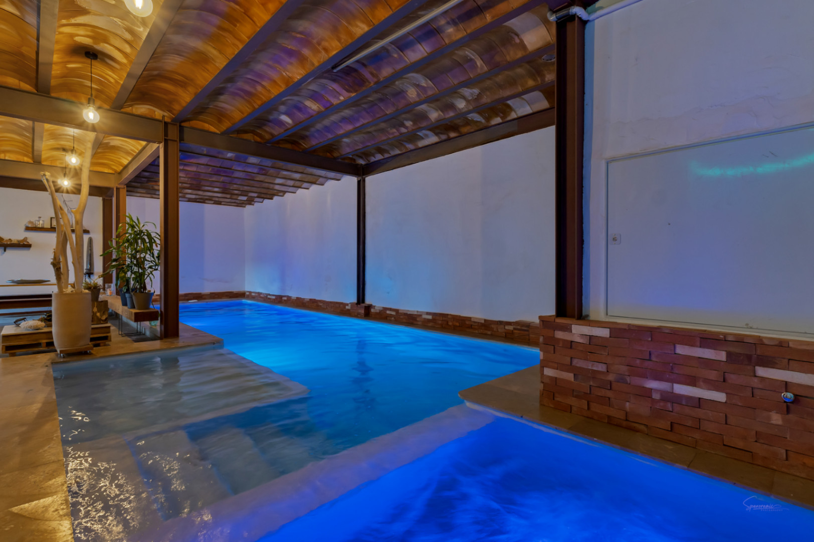 Verwarmd binnenzwembad met blauwe LED-verlichting, gewelfde terracotta plafonds en zichtbare stalen balken voor een rustieke en industriële uitstraling.