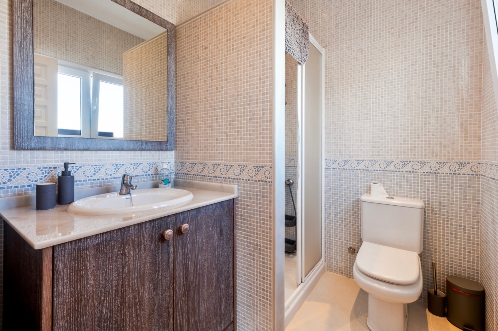 Badkamer met beige mozaïektegels, een blauwe decoratieve rand, donkerhouten wastafelmeubel met marmeren blad en een douchecabine.