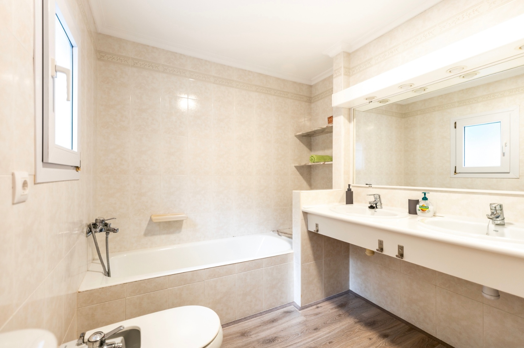 Badkamer voorzien van een ligbad, bidet en dubbele wastafel. De ruimte is afgewerkt met beige tegels, een grote verlichte spiegel en een vloer met houtlook.