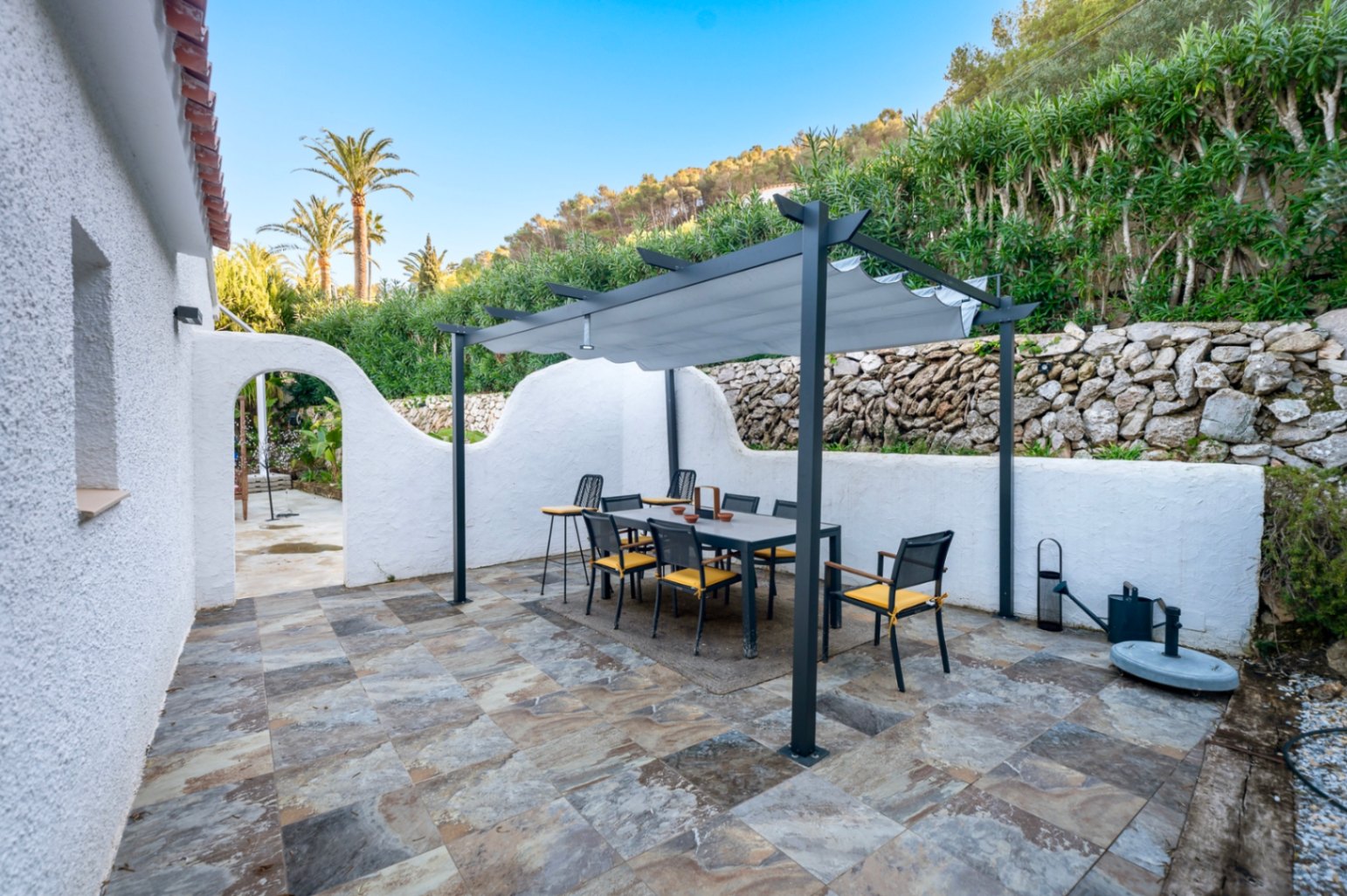 Buitenterras met natuurstenen tegels en een eethoek onder een moderne pergola. De ruimte wordt begrensd door droge stenen muren en mediterrane beplanting.