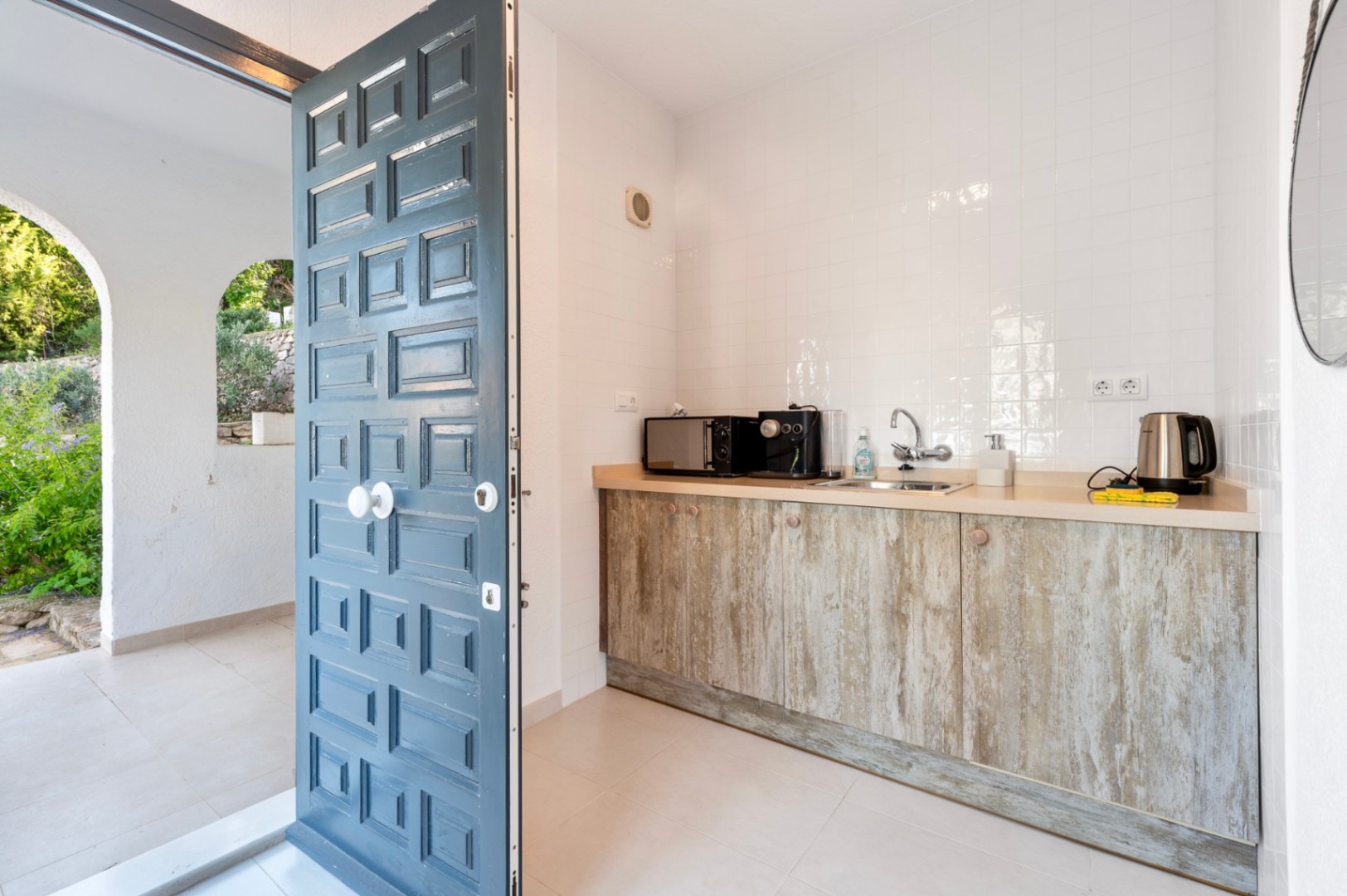 Compacte kitchenette met kasten in rustieke houtlook en een witte tegelwand, gelegen naast een traditionele blauwe paneeldeur die naar buiten leidt.