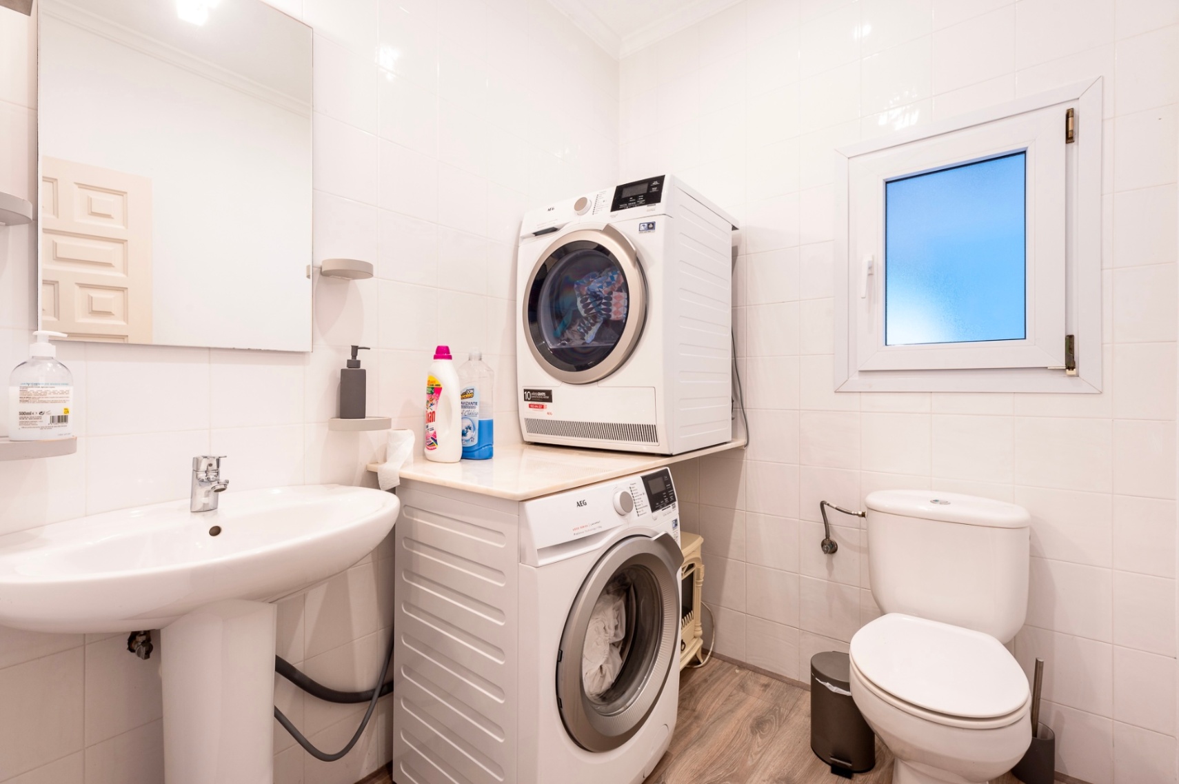 Functionele wasruimte en gastentoilet met wit betegelde wanden, uitgerust met AEG wasmachine en droger, een wastafel op zuil en vloer met houtlook.