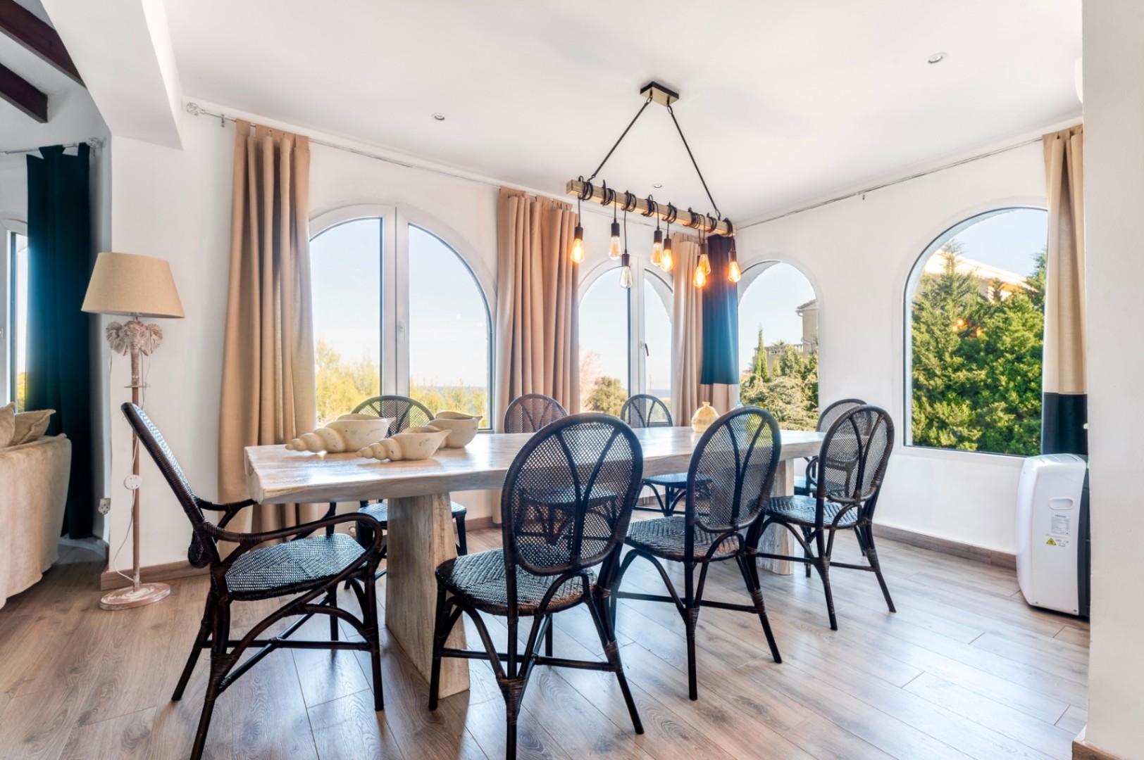 Lichte eetkamer met een rustieke houten tafel, zwarte rotan stoelen en karakteristieke boogvensters die uitkijken op de omliggende tuin.