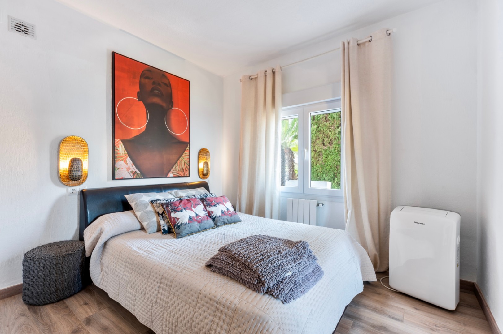 Lichte slaapkamer met houtlook vloer, donker lederen hoofdbord en opvallende wanddecoratie. Voorzien van gouden wandlampen, radiator en een mobiele airco.