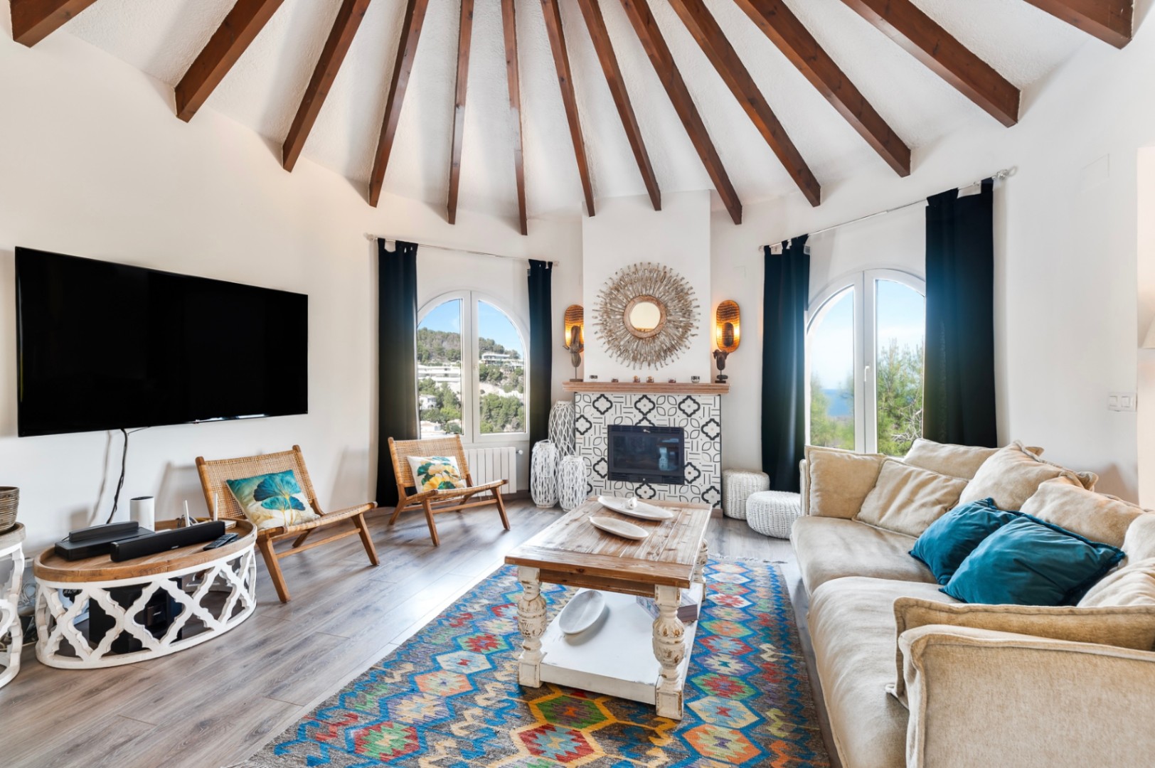 Ronde woonkamer met een gewelfd plafond met houten balken, een betegelde haard en boogvensters die uitkijken op het mediterrane landschap.