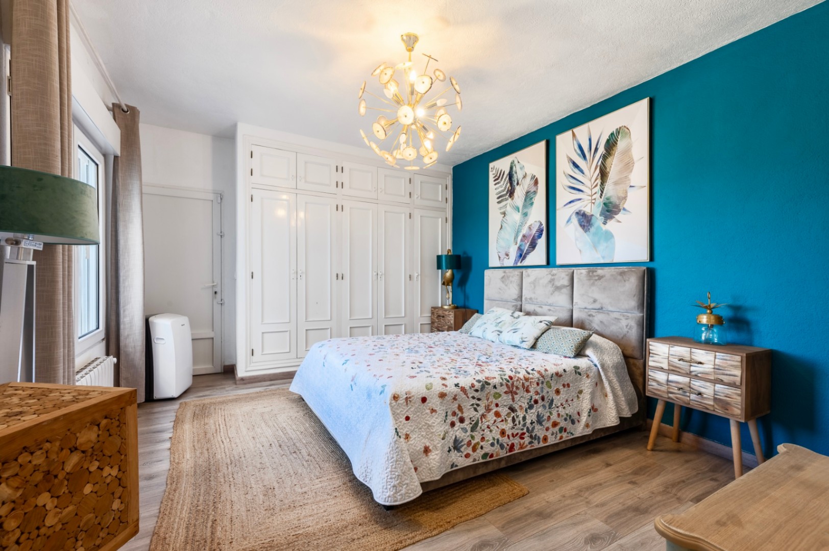 Slaapkamer met een blauwe accentmuur, witte inbouwkasten, houten vloer en een opvallende gouden designlamp boven het bed met gestoffeerd hoofdbord.