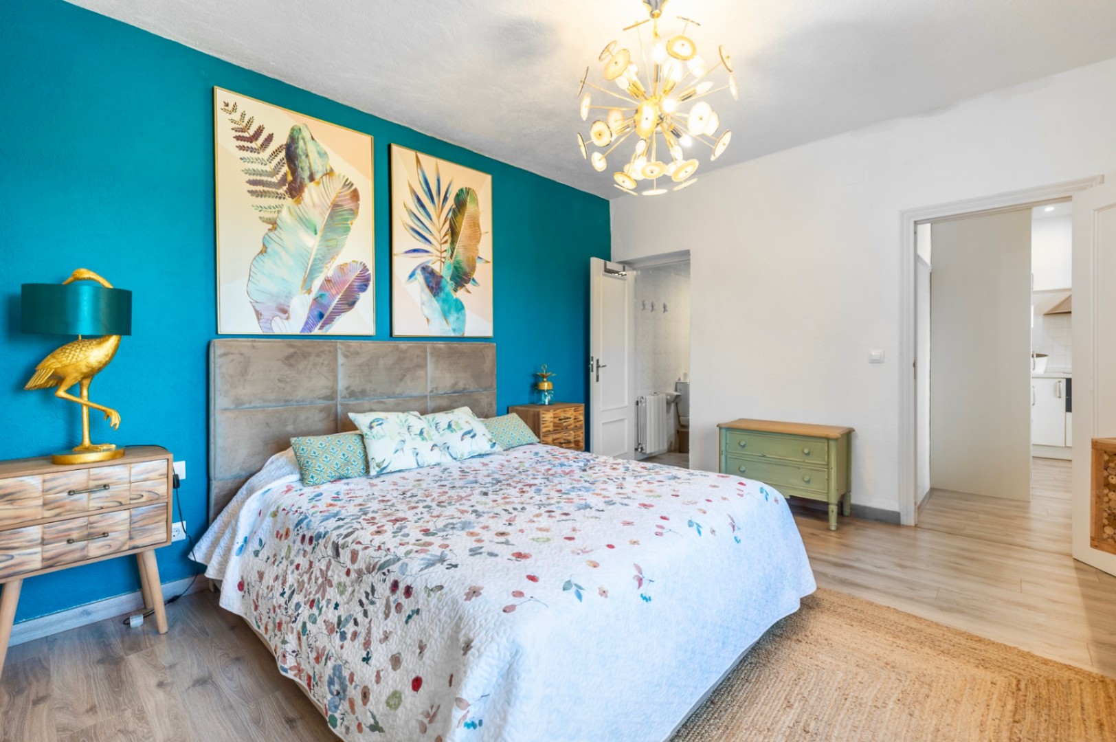 Slaapkamer met een turquoise accentmuur, botanische prints en een gouden kroonluchter. Inclusief en-suite badkamer en lichte laminaatvloer met jute tapijt.