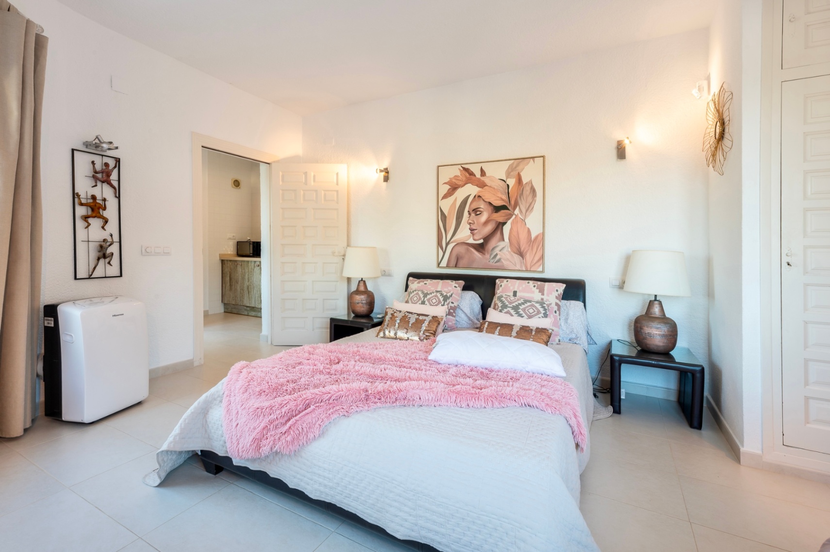 Slaapkamer met tweepersoonsbed, roze accenten en witte inbouwkasten. De ruimte is voorzien van lichte tegelvloeren, wandlampen en een mobiele airconditioningunit.