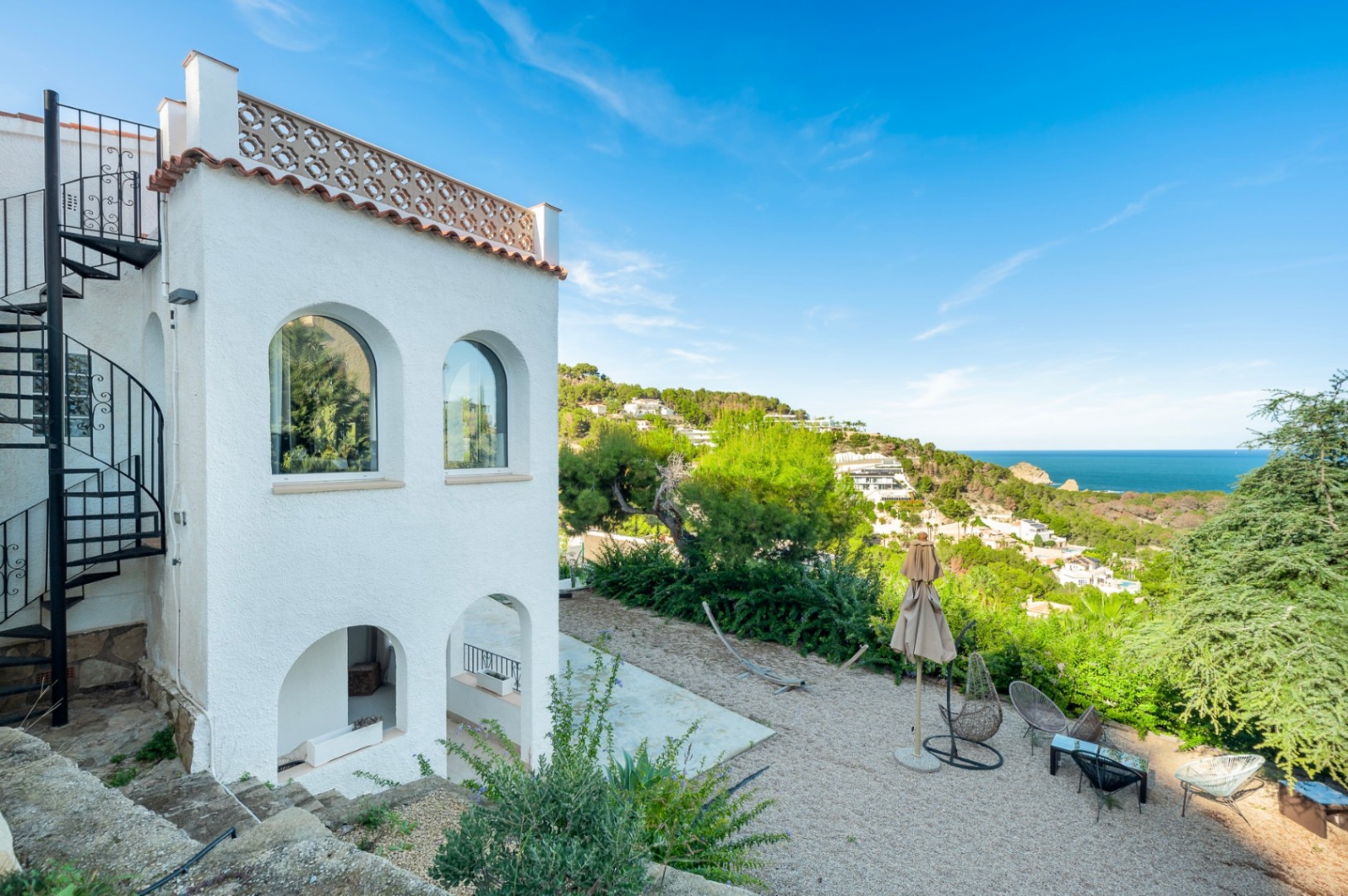 Witte mediterrane gevel met boogramen, een zwarte wenteltrap en een grindterras met zitgedeelte en panoramisch uitzicht over de kustlijn en de zee.