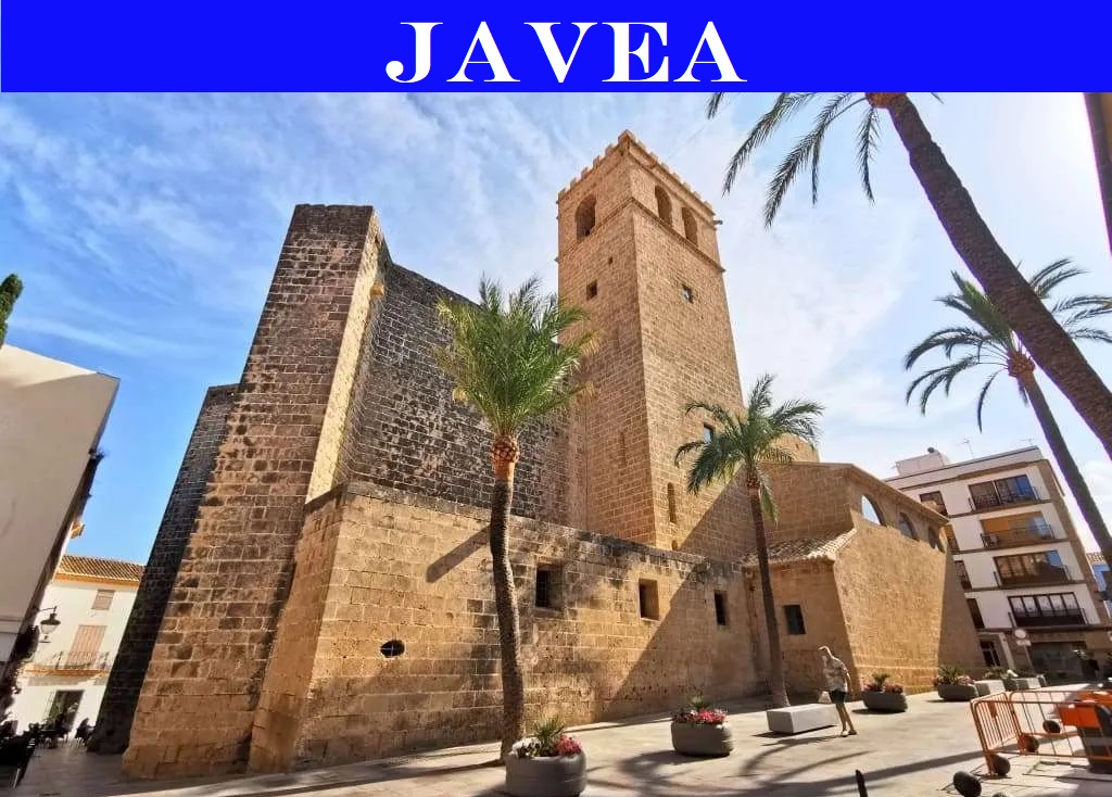 Buitenaanzicht van de historische versterkte kerk van San Bartolomé in Jávea, opgetrokken uit toscasteen met een karakteristieke klokkentoren en omringd door palmbomen.