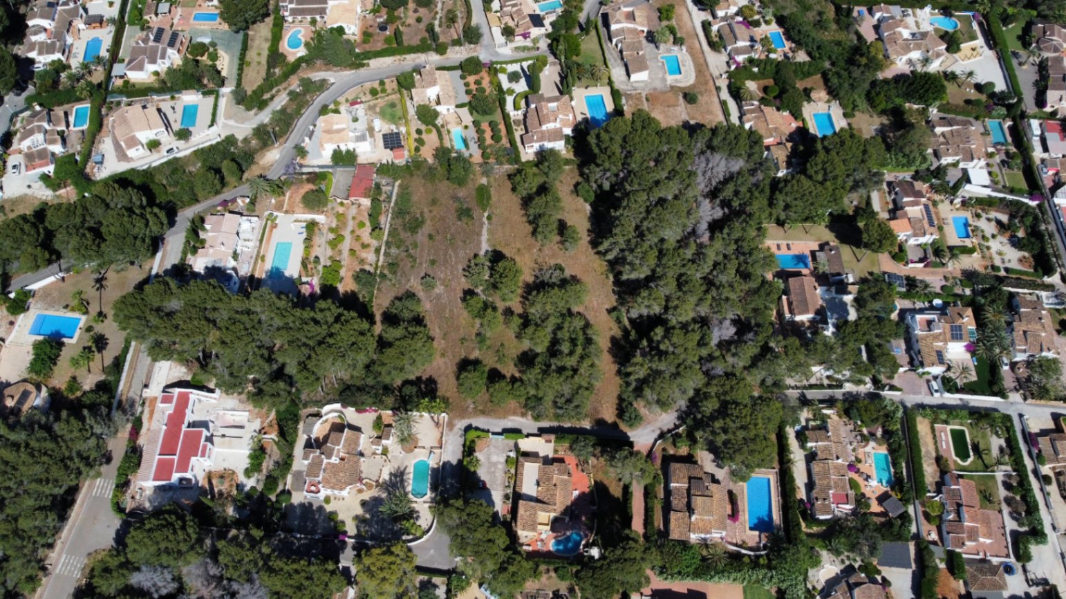Luchtfoto van het grote vlakke perceel met pijnbomen, gelegen in een volgroeide woonwijk en omringd door villa's met privézwembaden.