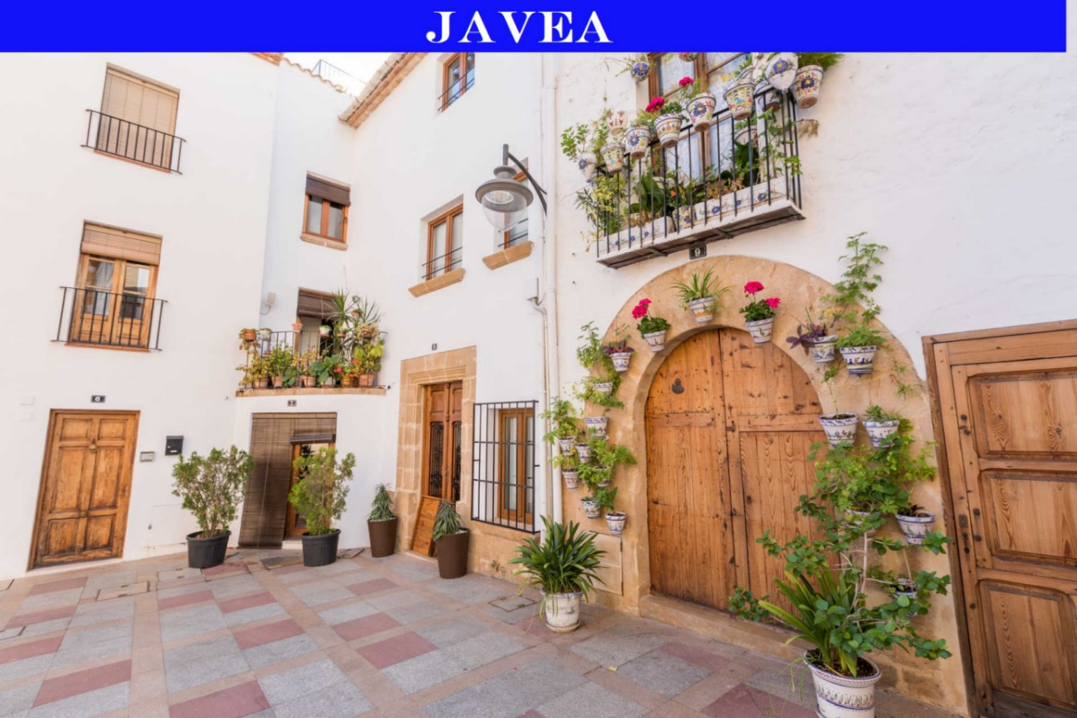 Traditionele mediterrane gevel in Jávea met houten deuren, een stenen boog en balkons versierd met keramische bloempotten en weelderige planten.