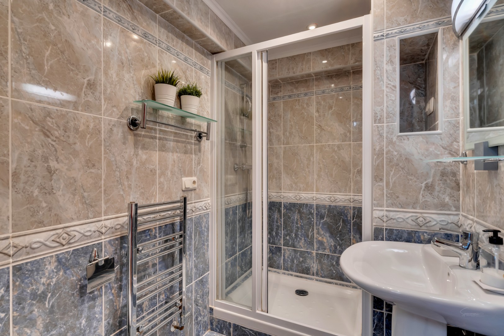 Badkamer met inloopdouche voorzien van wit frame, beige en blauwe marmerlook tegels, chromen handdoekradiator en een wastafel.
