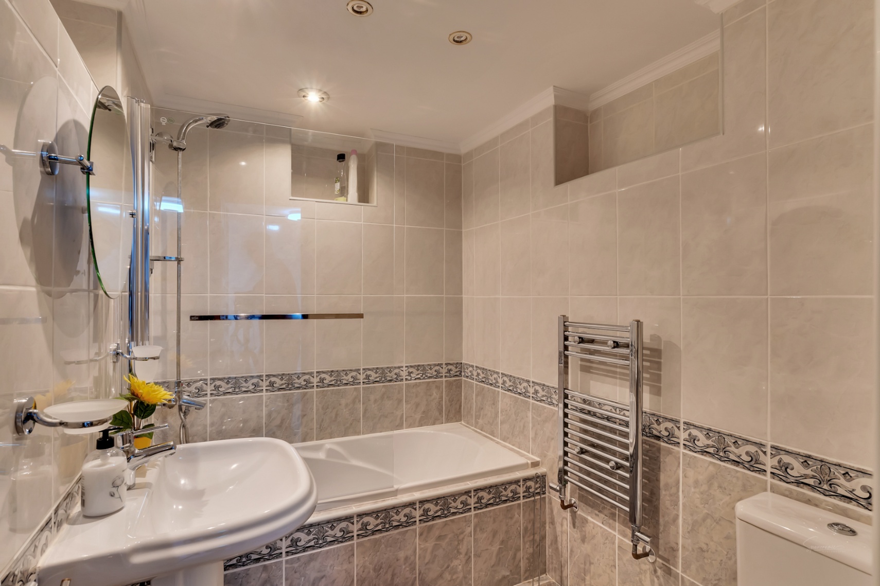 Badkamer met ligbad en glazen wand, crèmekleurige tegels met decoratieve rand, chromen handdoekradiator en inbouwspots in het plafond.