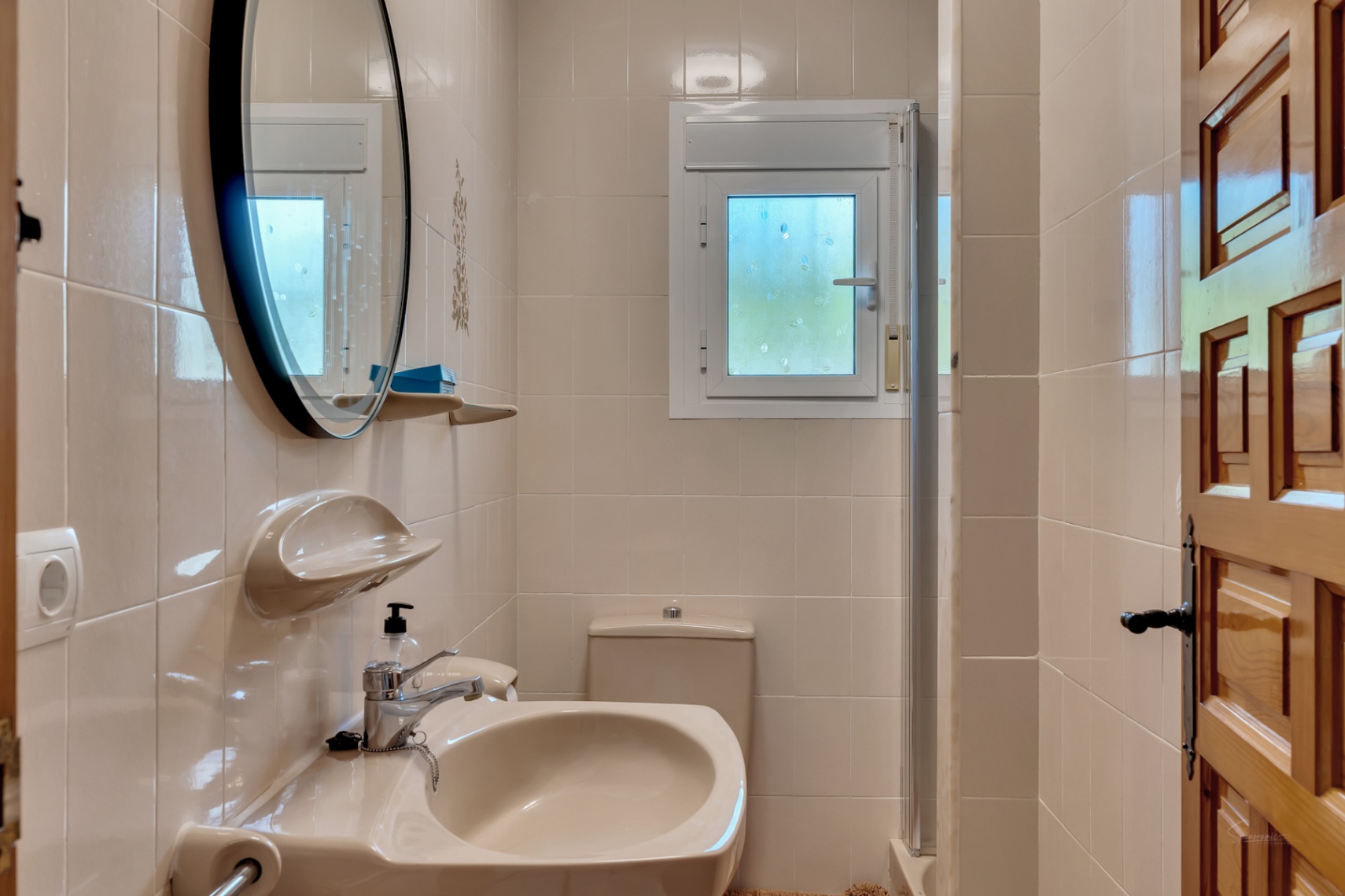 Functionele badkamer met crèmekleurige tegels, een ronde spiegel met zwart frame, keramische wastafel en een klein raam voor natuurlijke ventilatie.