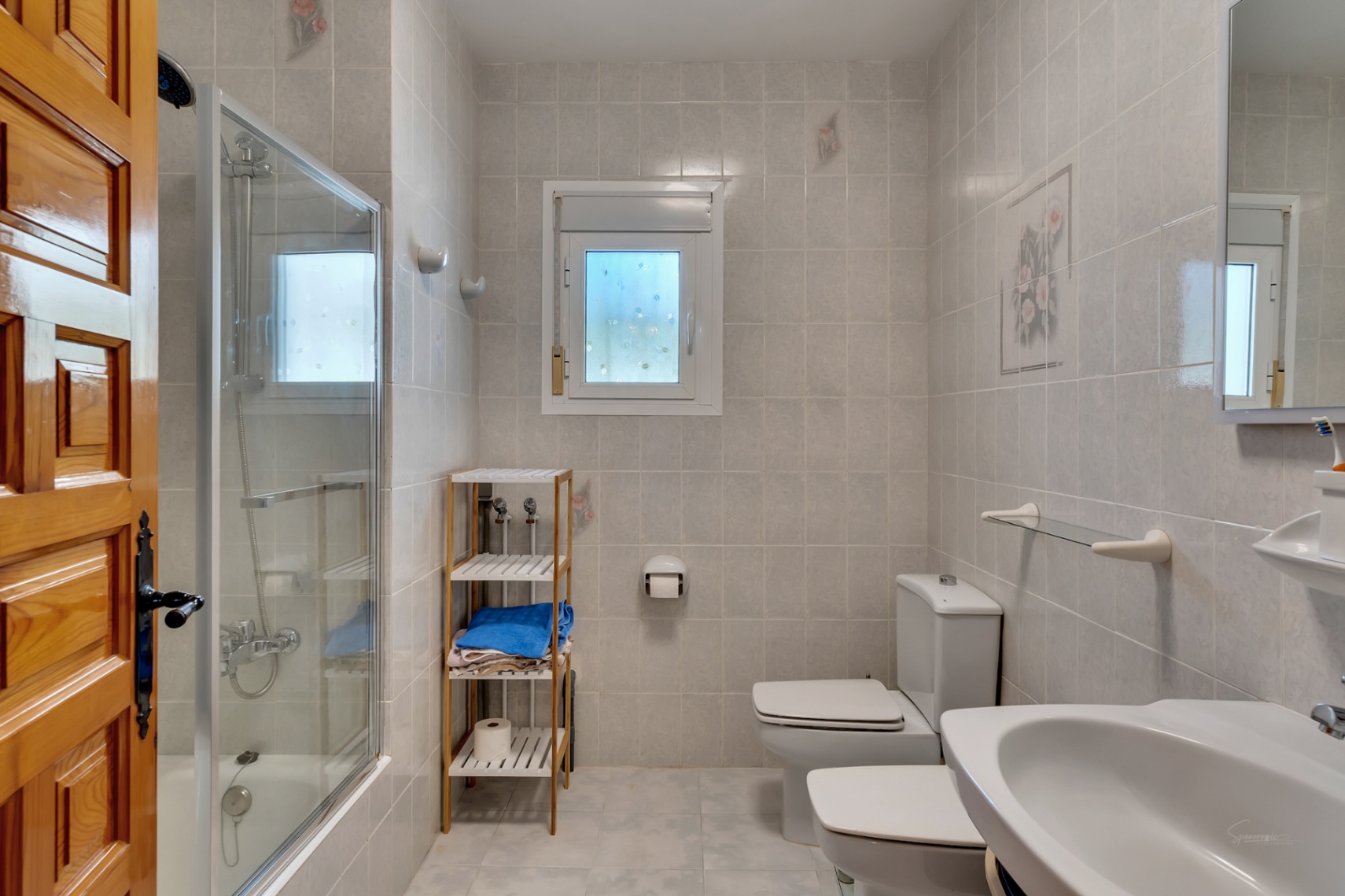 Functionele badkamer met glazen douchecabine, wit sanitair en betegelde wanden. Voorzien van een raam voor natuurlijk licht en een massief houten deur.