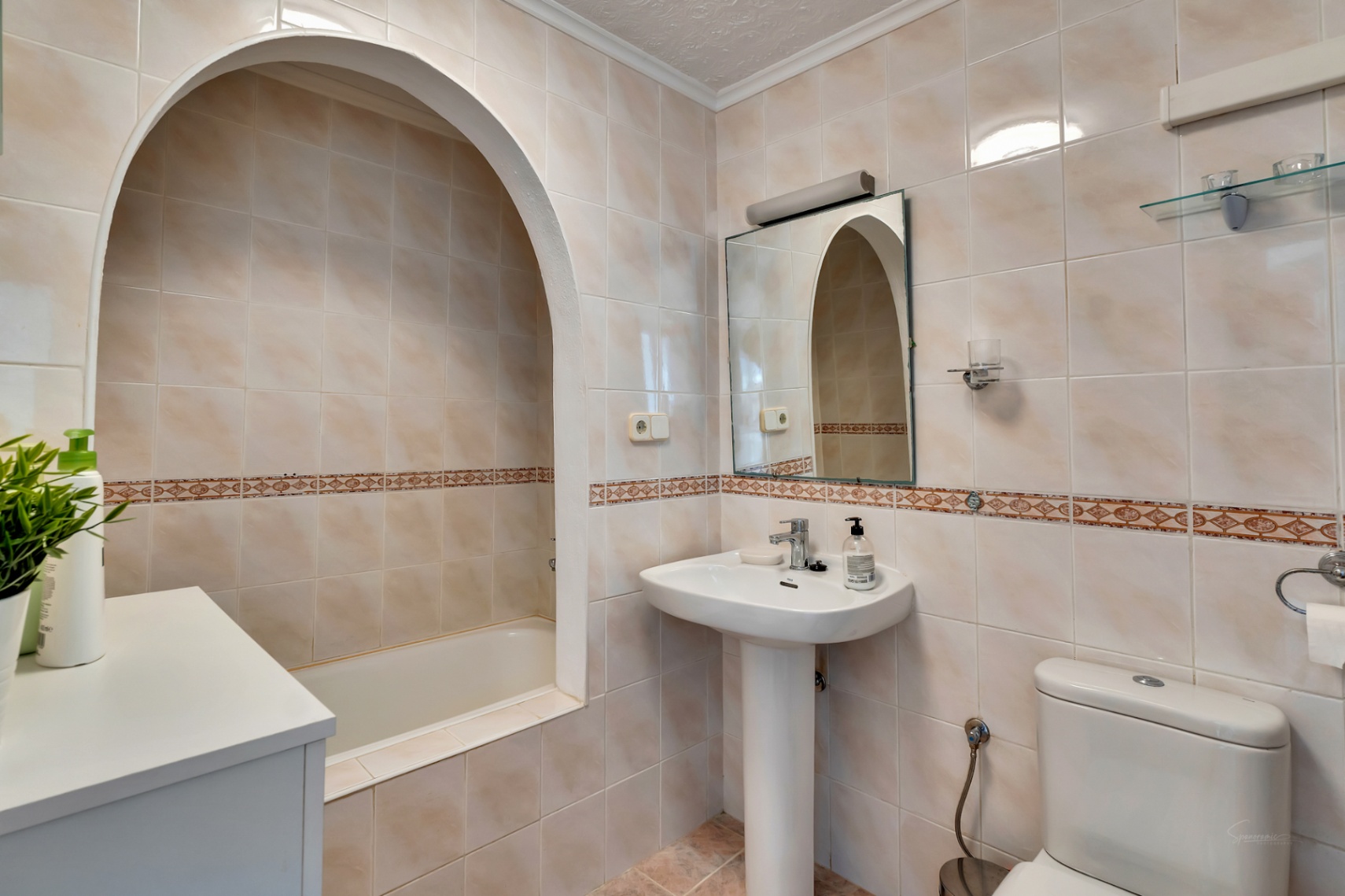 Klassieke badkamer met een ligbad in een karakteristieke boognis, een kolomwastafel en beige wandtegels met een decoratieve sierstrip.